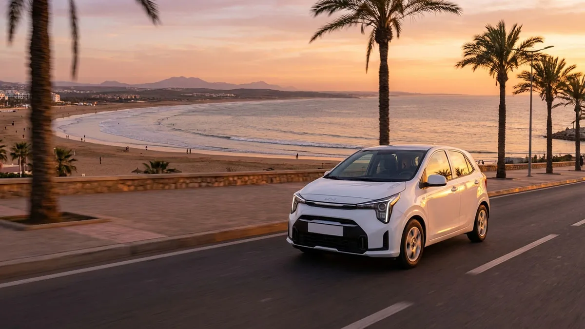 Kia Picanto 2025 roulant le long de la Corniche d’Agadir avec la mer et le ciel orangé en arrière-plan.