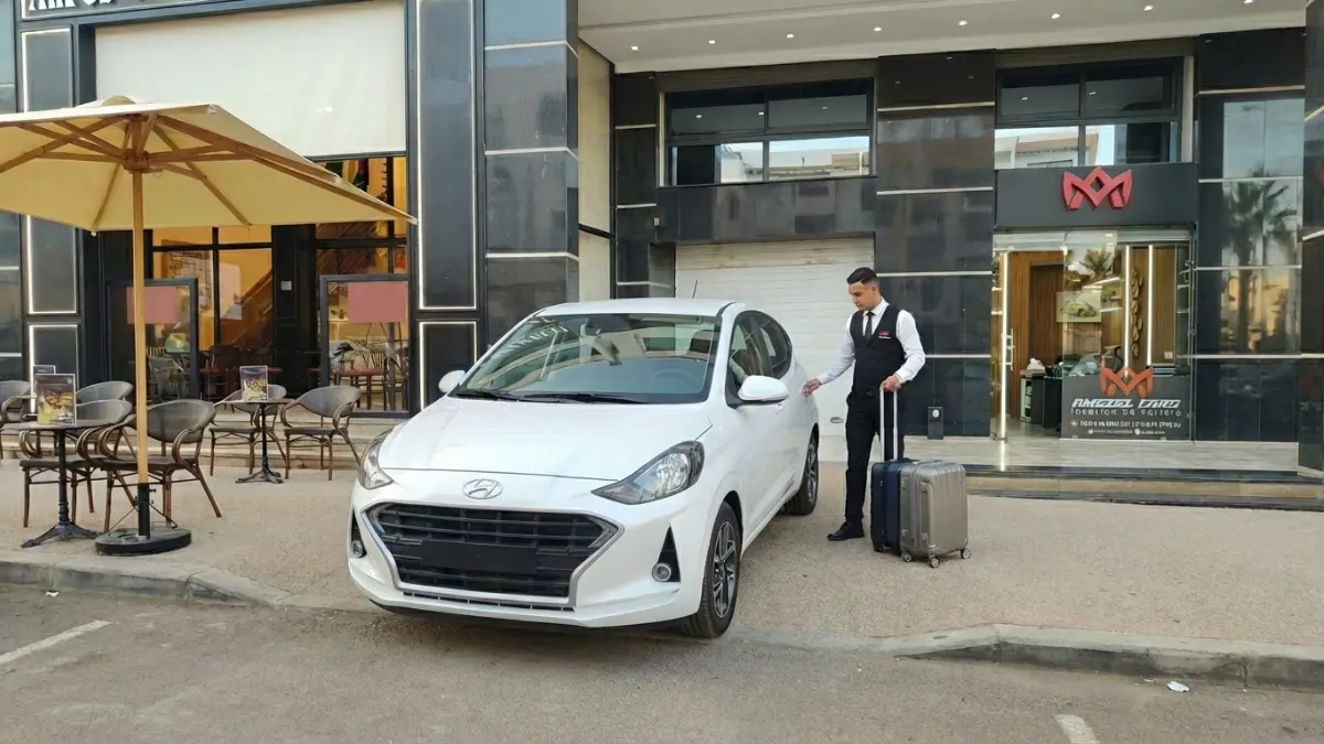 Une Hyundai i10 garée devant le comptoir Amseelcars à l’aéroport d’Agadir avec un agent souriant et bagages, Hyundai i10.
