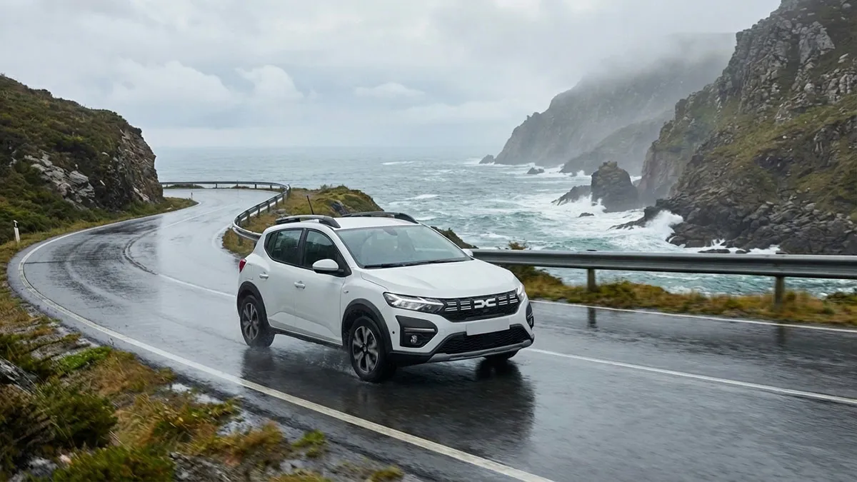 Dacia Sandero Stepway prenant un virage sur une route côtière marocaine humide avec falaises et océan en arrière-plan, sans texte dacia sandero Stepway