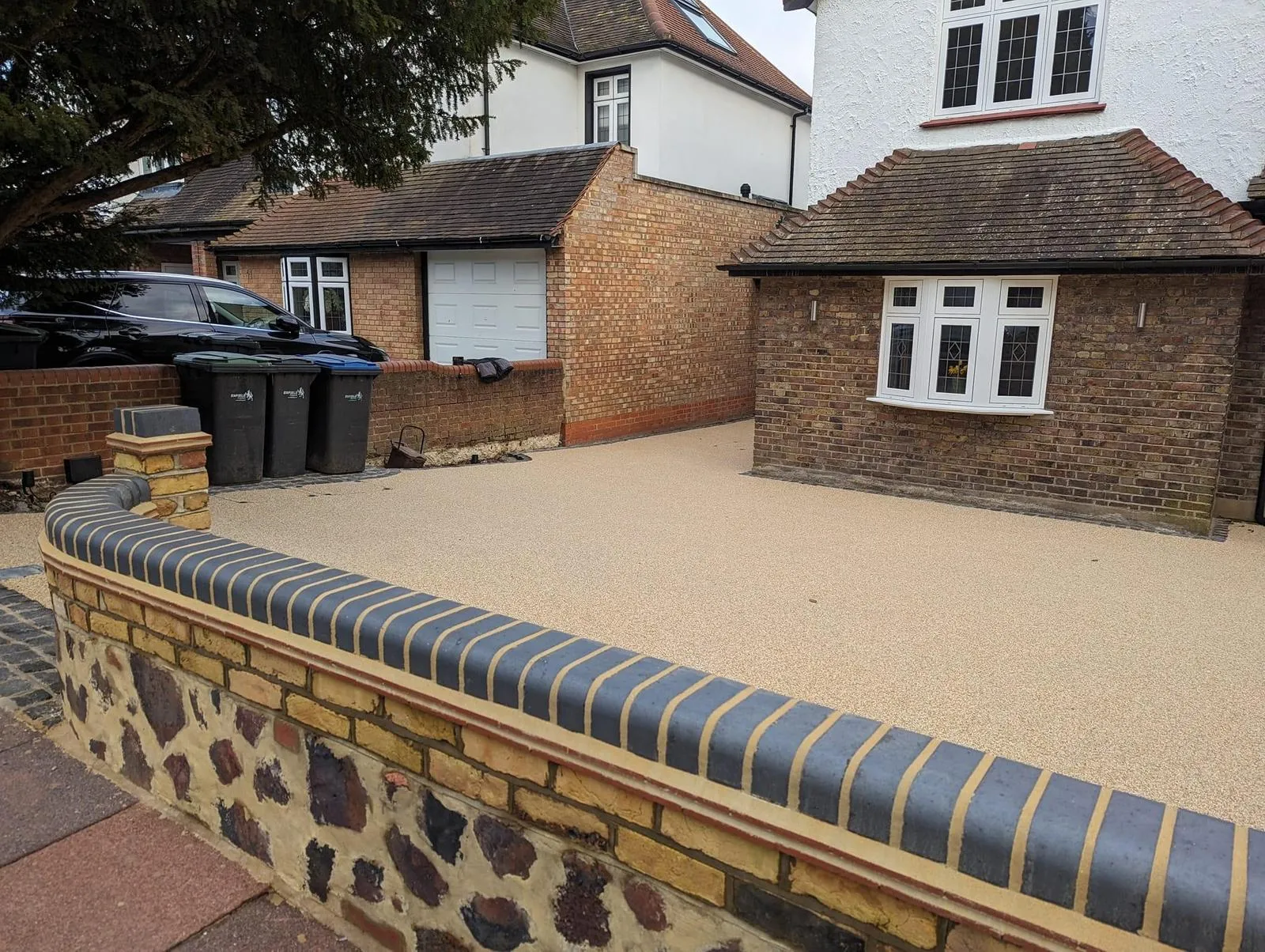 A modern, light-coloured resin-bound driveway with a curved brick wall and a house in the background.