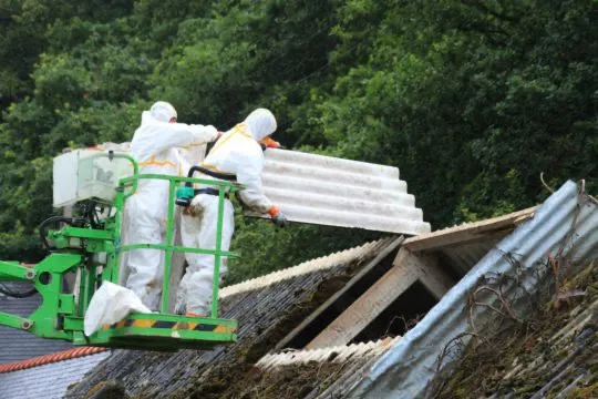 Professionals in white hazmat suits using a MEWP to remove corrugated asbestos roofing from an old building.