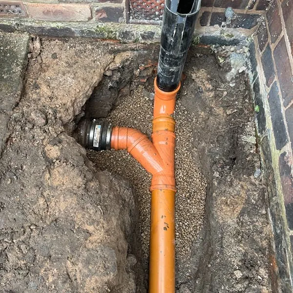 An open trench exposing orange drainage pipes with a Y-junction and a black downpipe, set against a brick wall, indicating plumbing work.