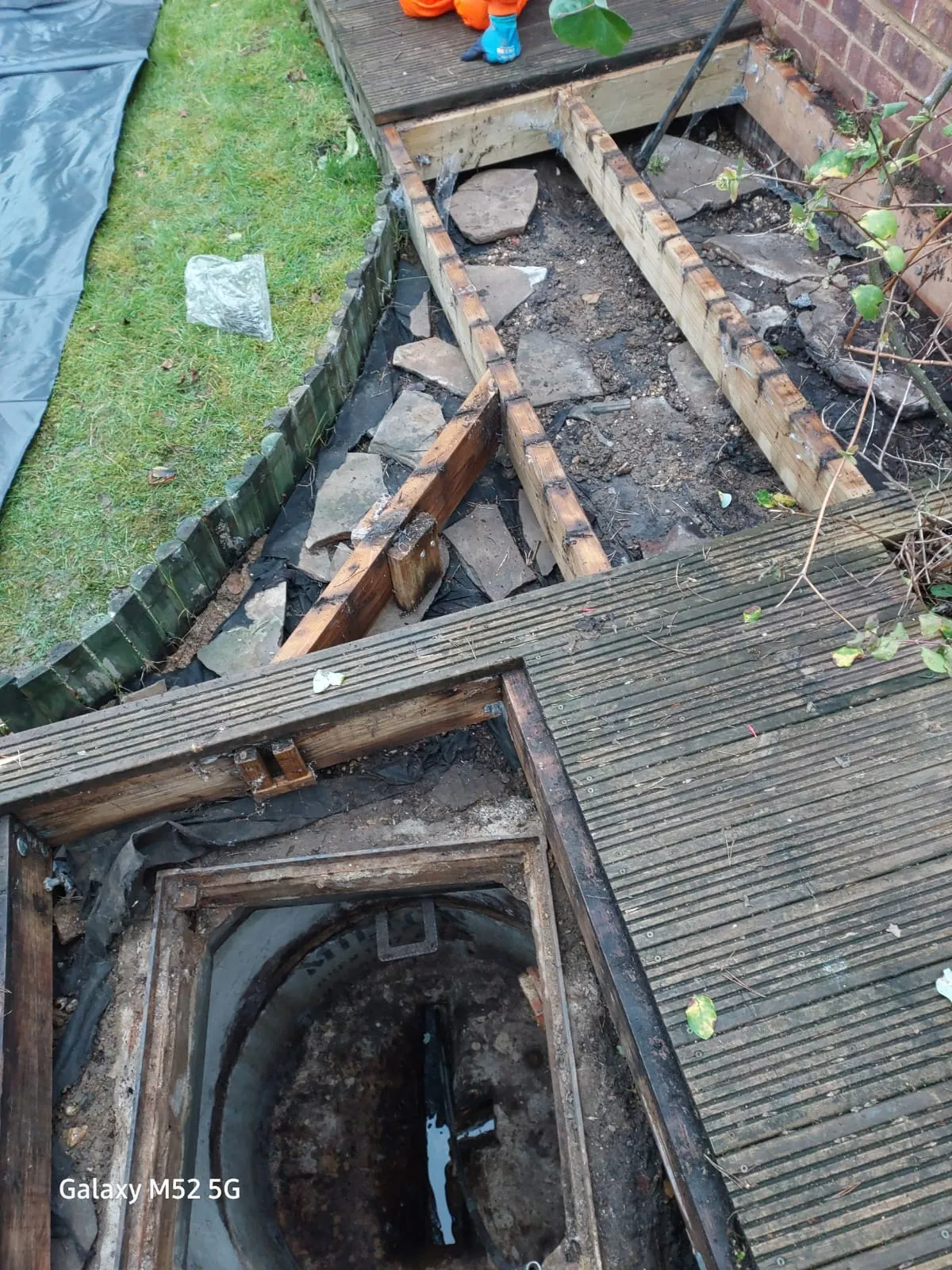 An open drain manhole with a small amount of water visible, surrounded by dismantled wooden decking, soil, and grass, indicating ongoing drainage work.