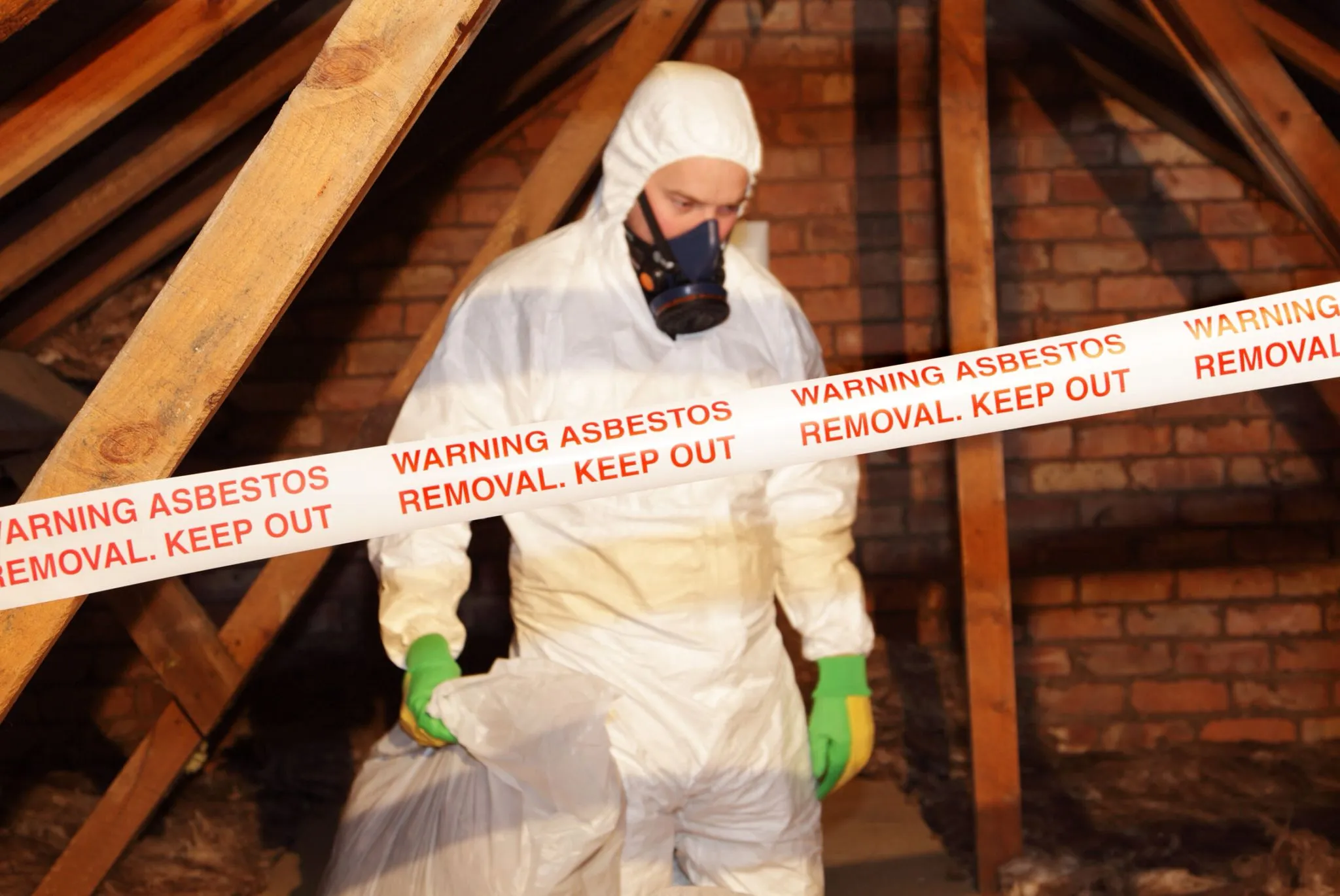Professional asbestos removal in an attic, showing a worker in a protective suit and respirator with warning tape.