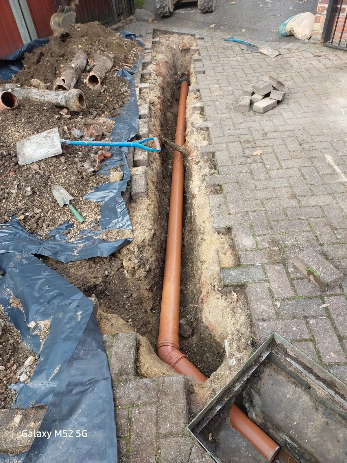 An excavated trench showing new orange drainage pipes laid, with old pipes and soil piled up, beside a paved surface.