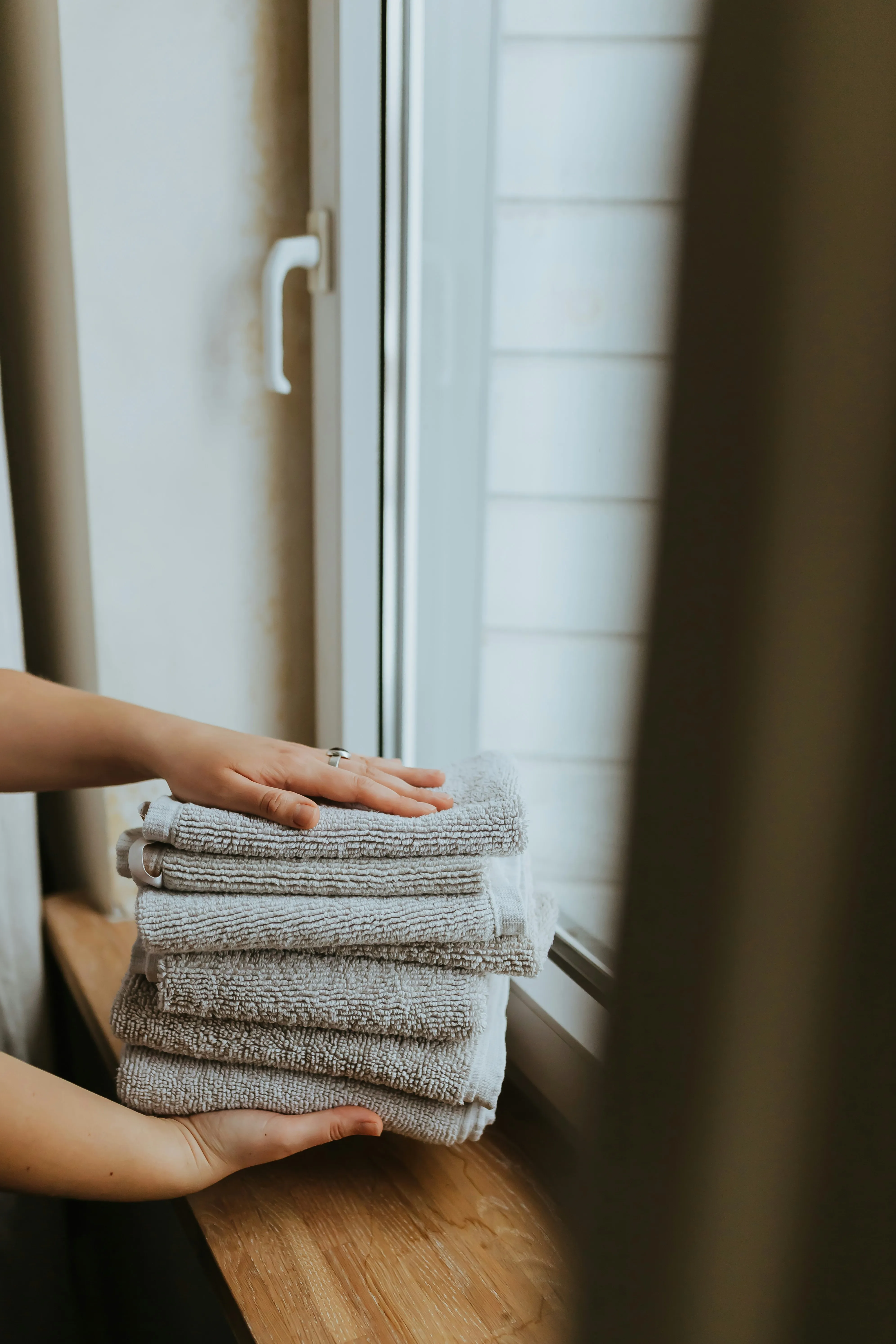 Hands carefully stacking clean, folded grey towels on a wooden surface by a window, ready for use.
