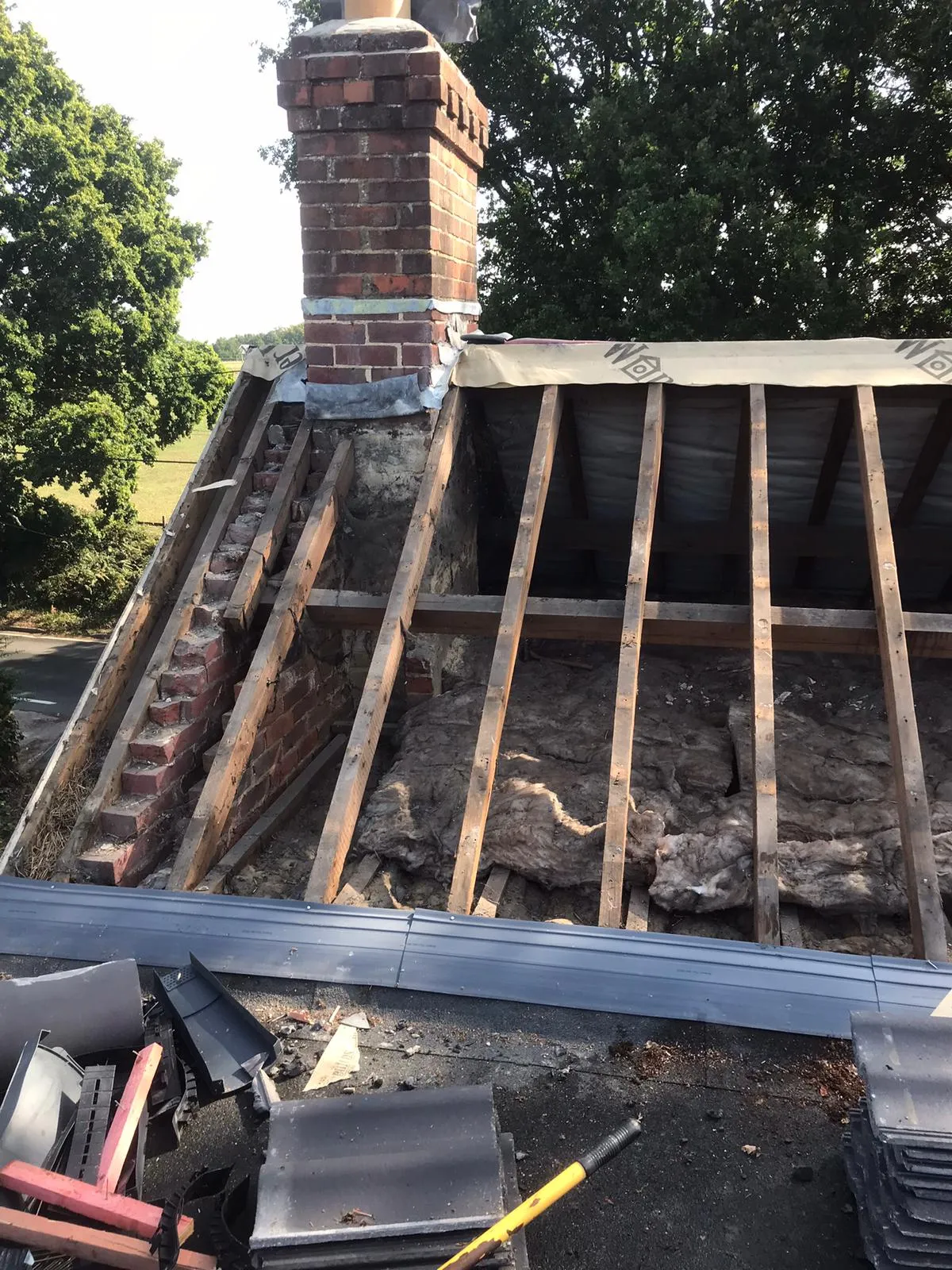 Roof repair in progress on a residential property, showing a brick chimney with new lead work, exposed wooden roof structure, insulation, and new tiles.