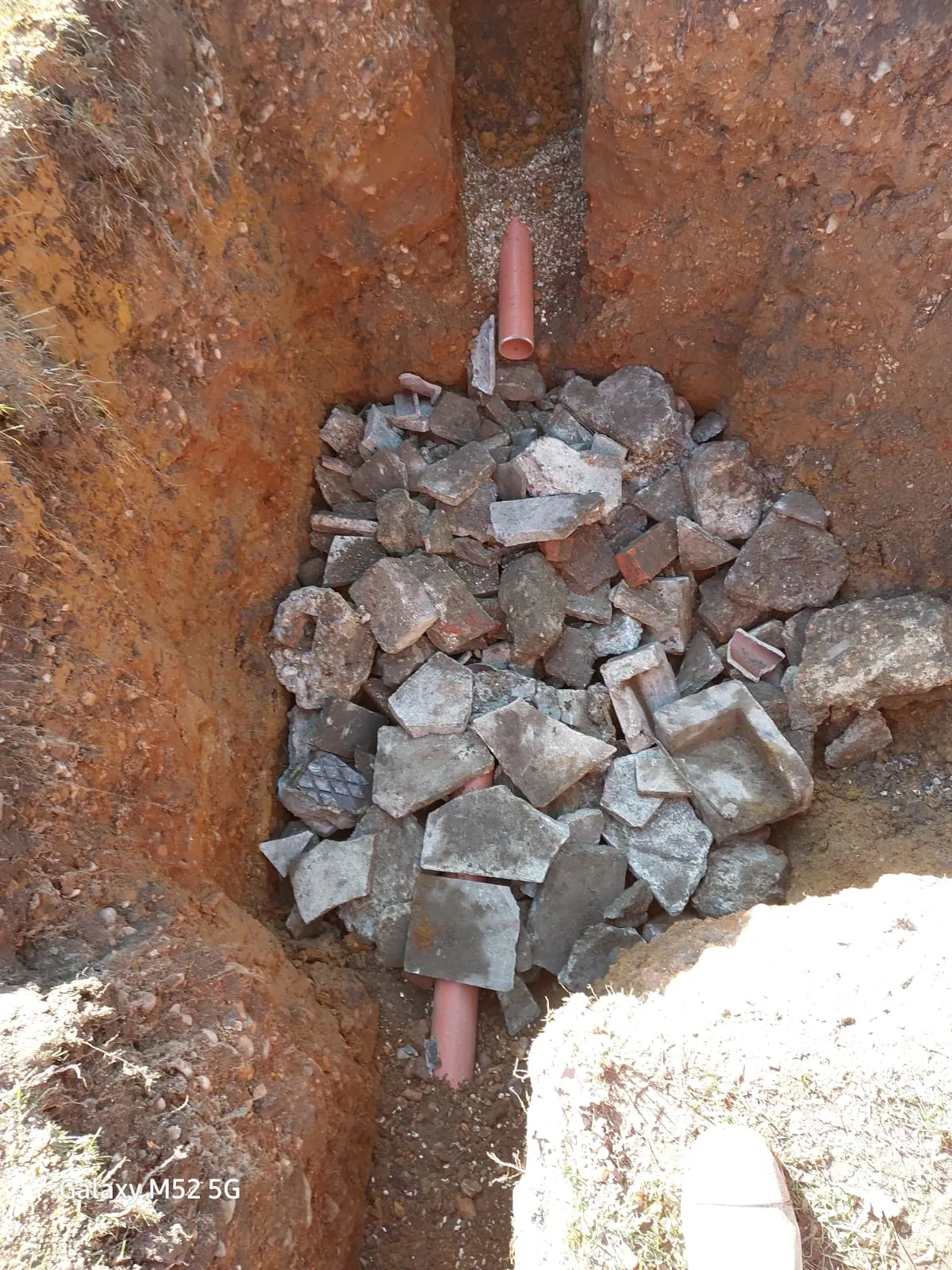 A deep trench dug in the earth, showing sections of orange plastic drainage pipes partially buried among broken concrete pieces and soil.