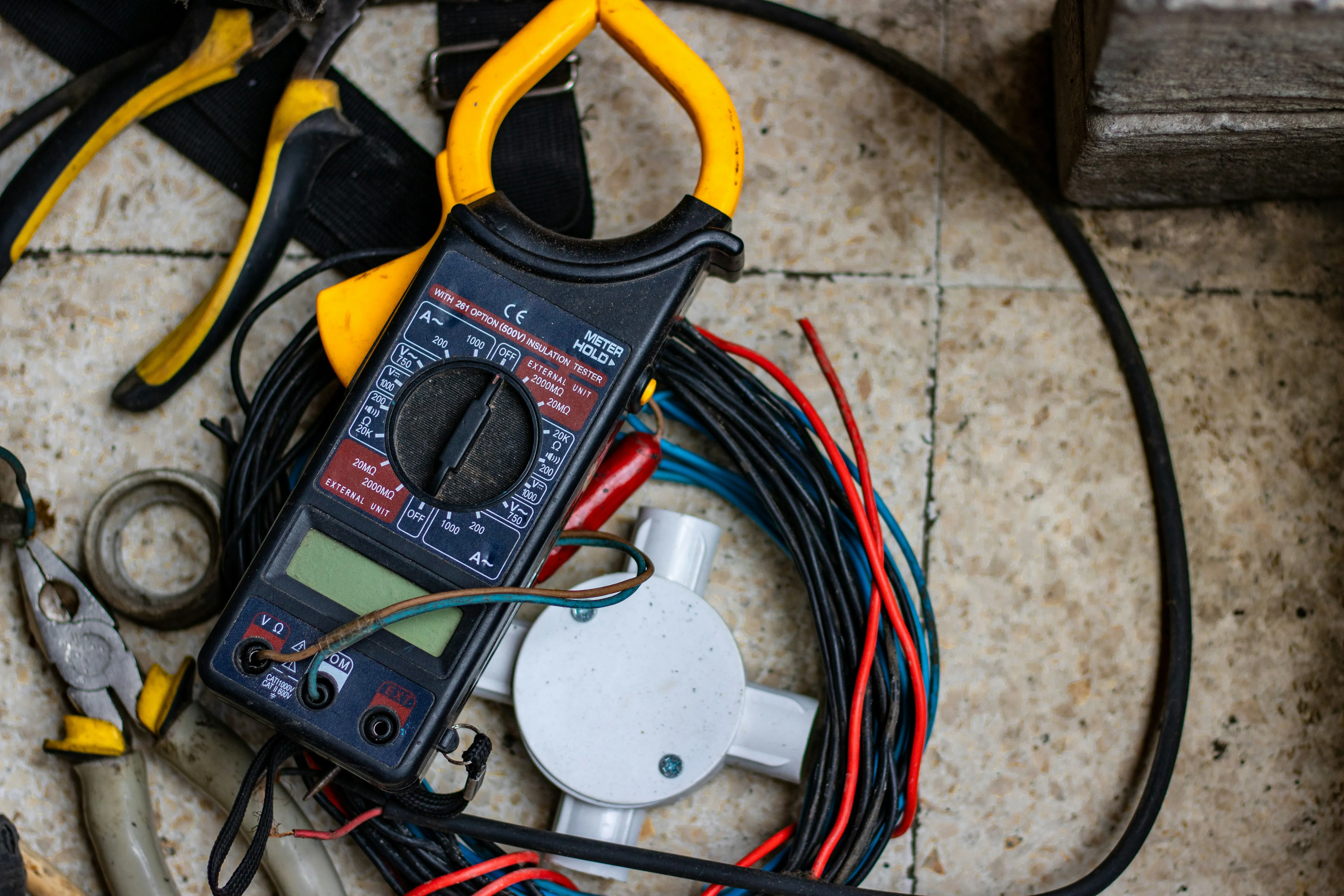 Electrician's tools and electrical components, including a clamp meter, wires, and junction box, on a tiled floor.