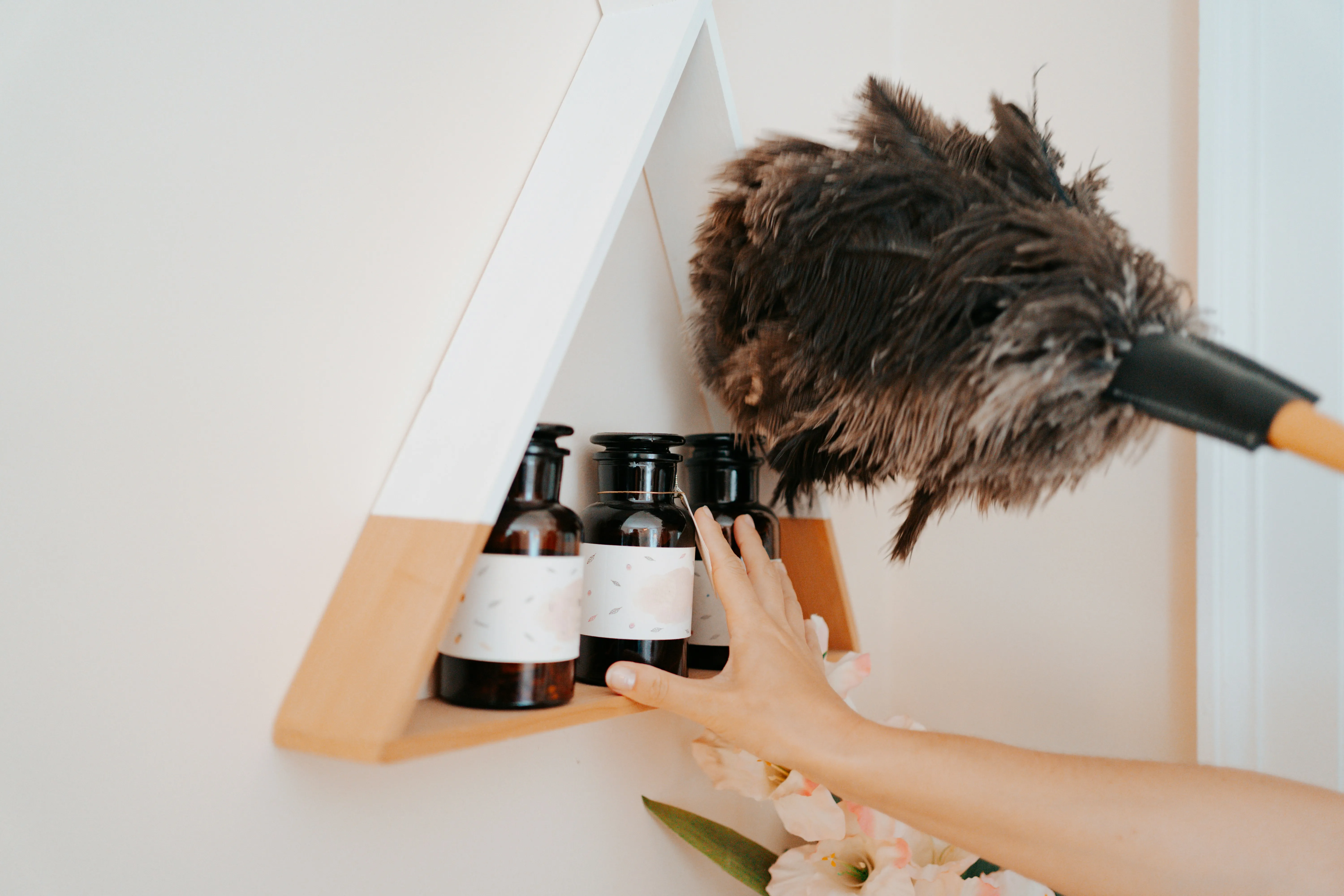 A hand using a feather duster to clean a decorative shelf with bottles, illustrating domestic cleaning.