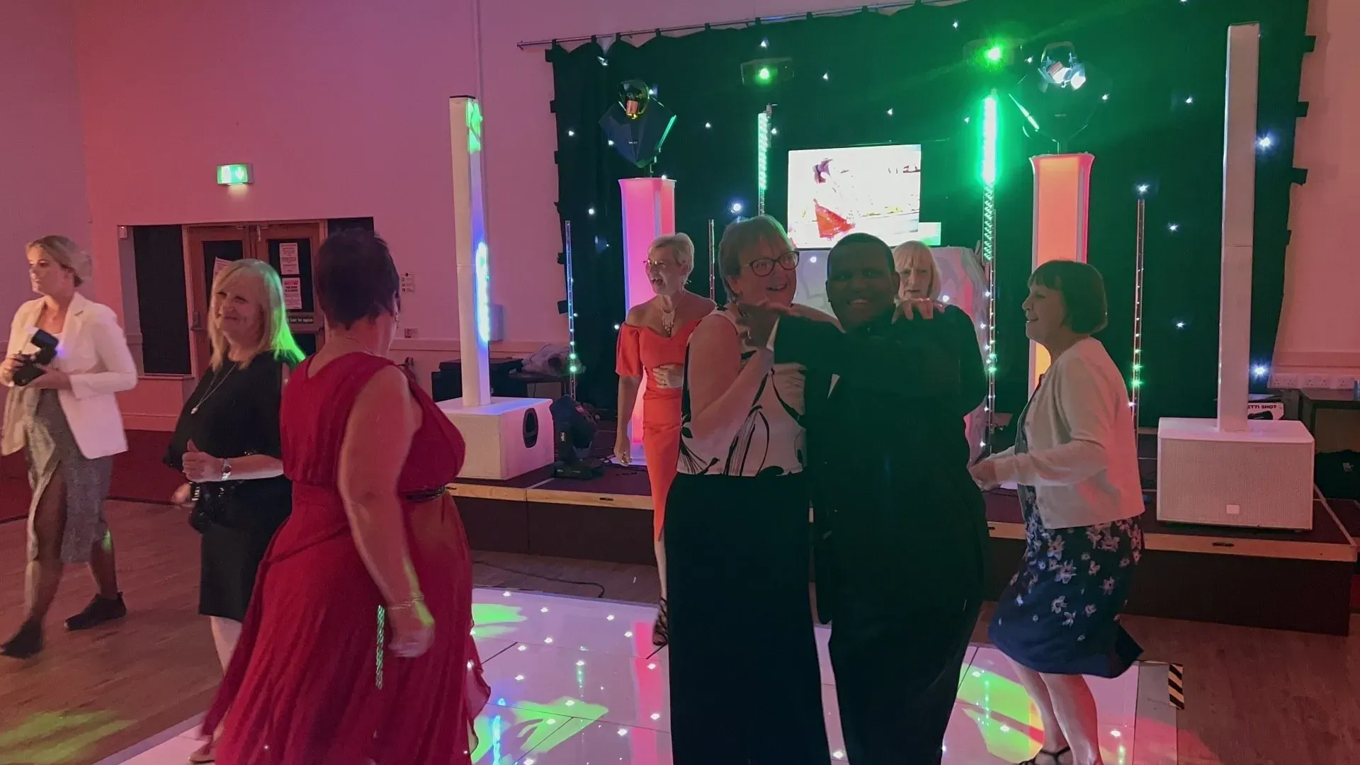 A group of happy guests dancing on a white LED starlit dancefloor, with colourful green and pink disco lights, large speakers, and an LED screen in the background at an event.