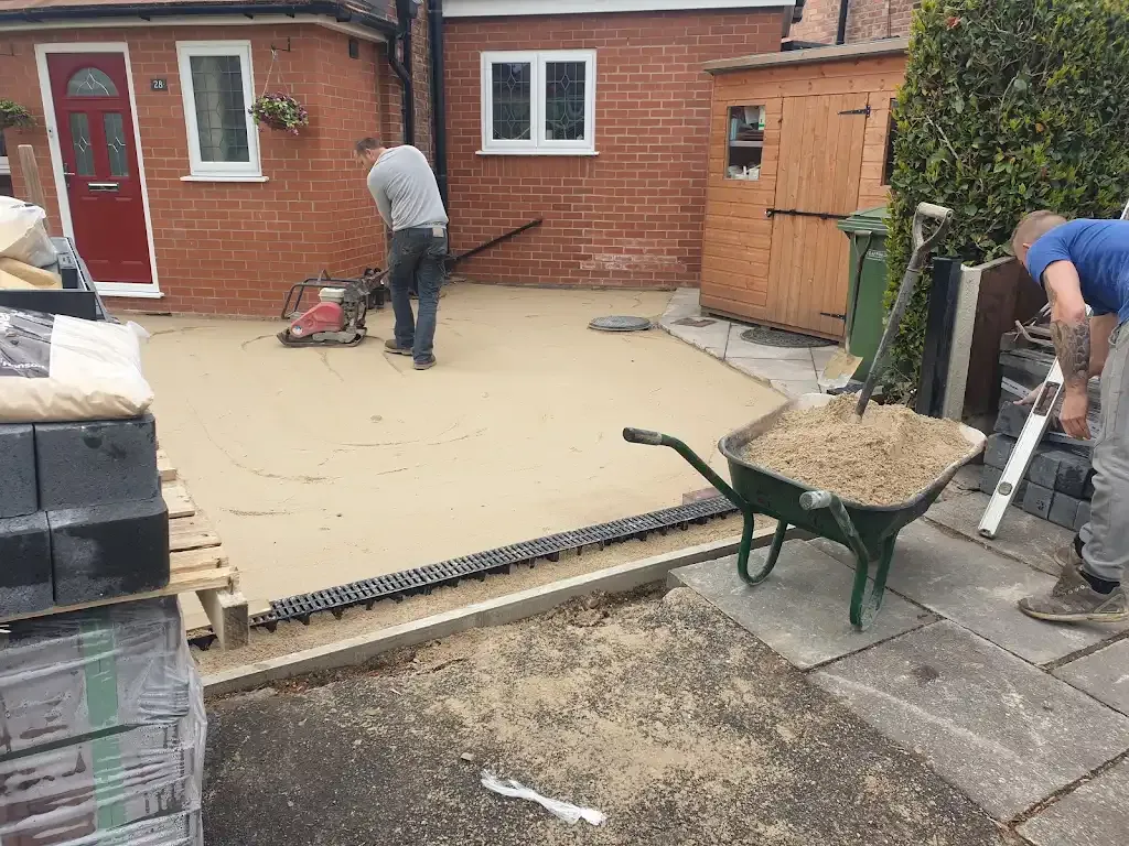Professional groundwork and preparation for a new block paving driveway installation, showing workers using a wacker plate and wheelbarrow.