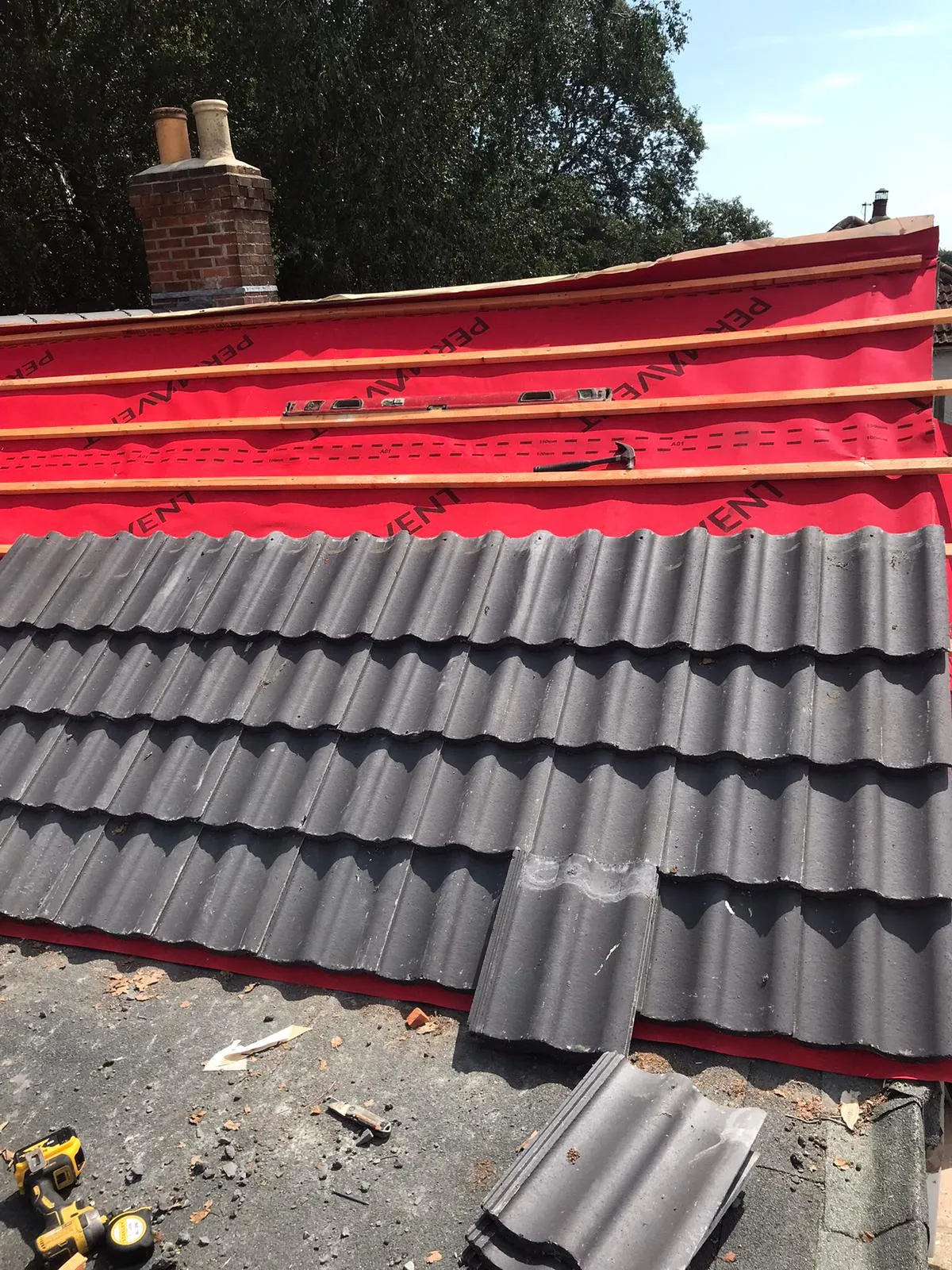 Professional installation of dark grey concrete roof tiles on a pitched roof, revealing red underlayment, wooden battens, and a brick chimney.