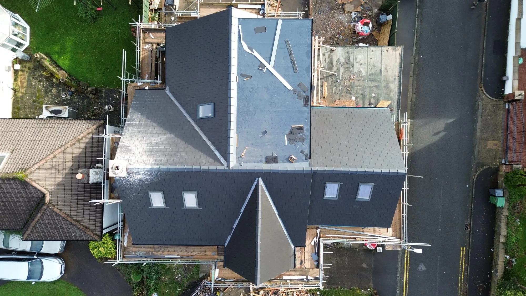 Drone view of a house during a roof replacement, showing new dark grey tiled sections, a flat roof in progress, Velux windows, and scaffolding around the building.