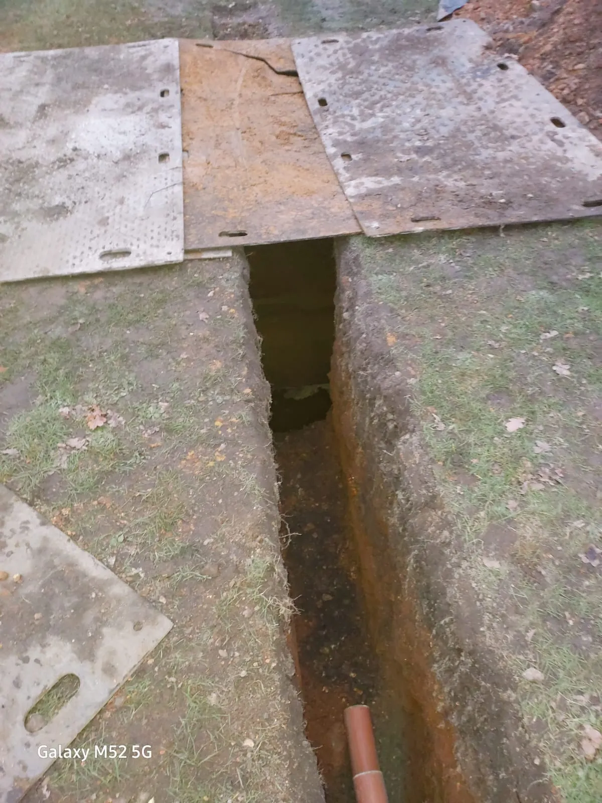 An excavated trench showing a newly laid brown drainage pipe during an installation or repair project.