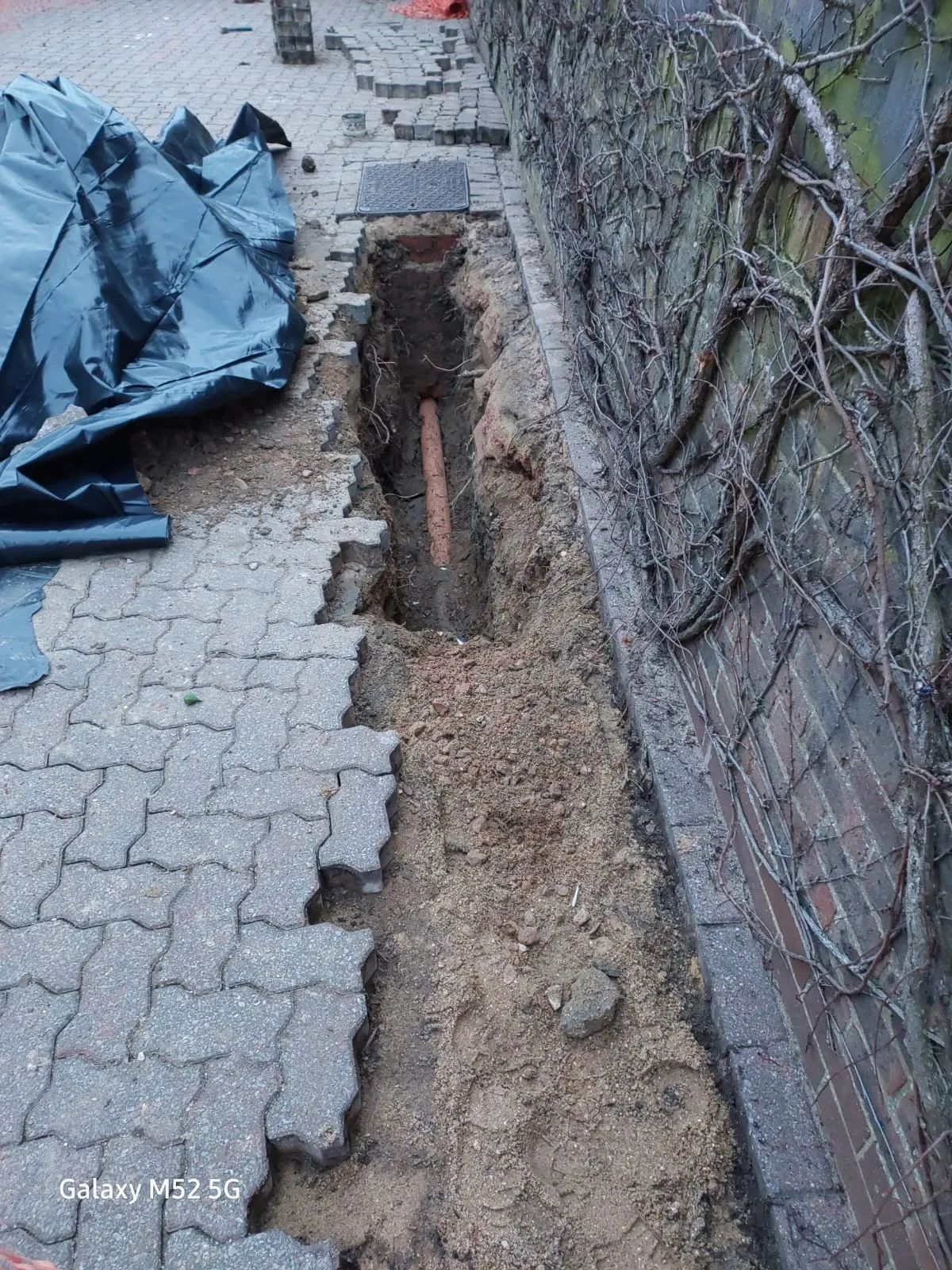 Excavation site for drain repair, showing an exposed pipe under removed paving stones next to a brick wall.