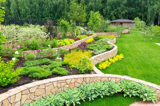 A landscaped garden featuring curved stone retaining walls, vibrant flower beds, and a lush green lawn.