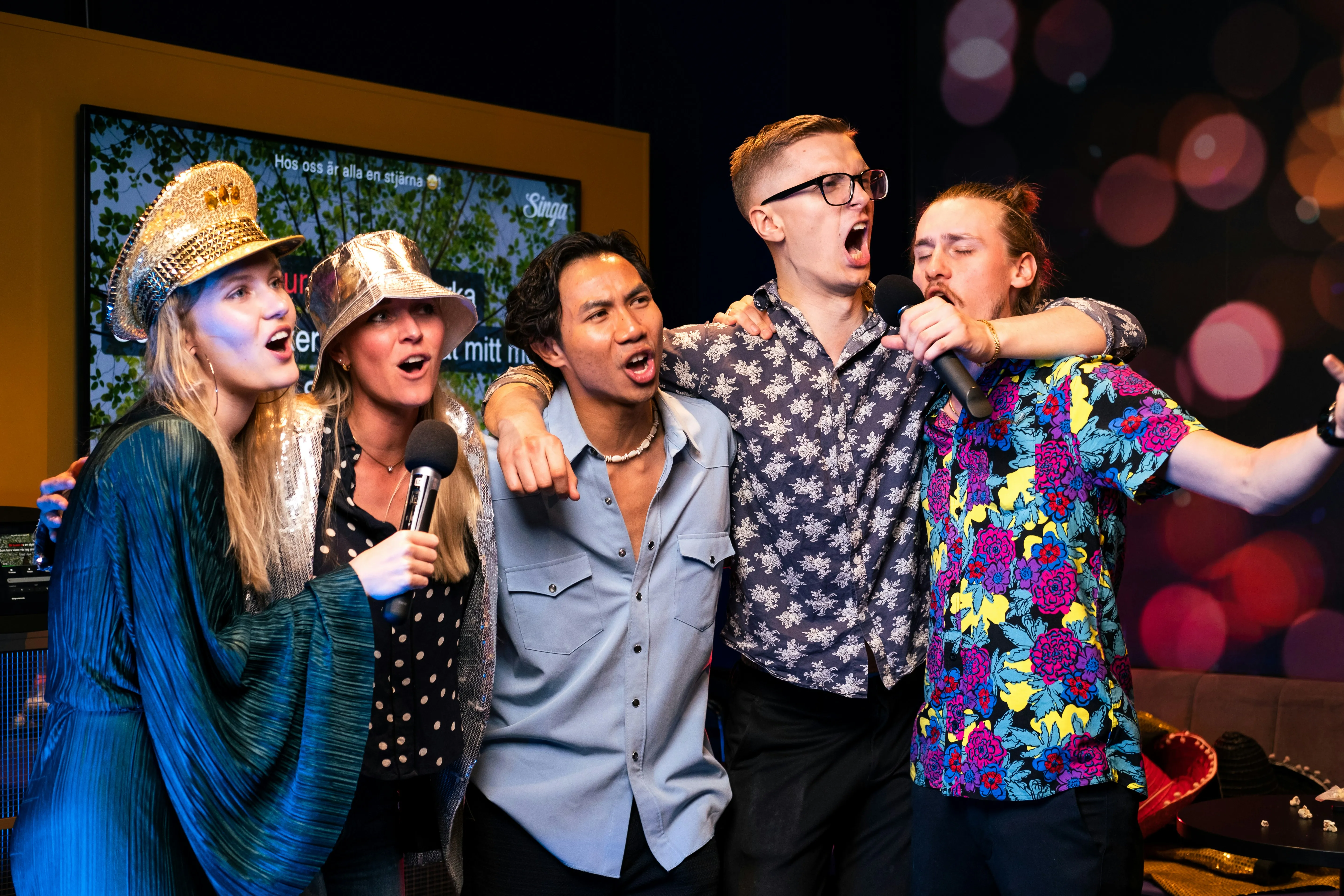 Group of friends enthusiastically singing karaoke together, some wearing party hats, with microphones and a screen in the background.