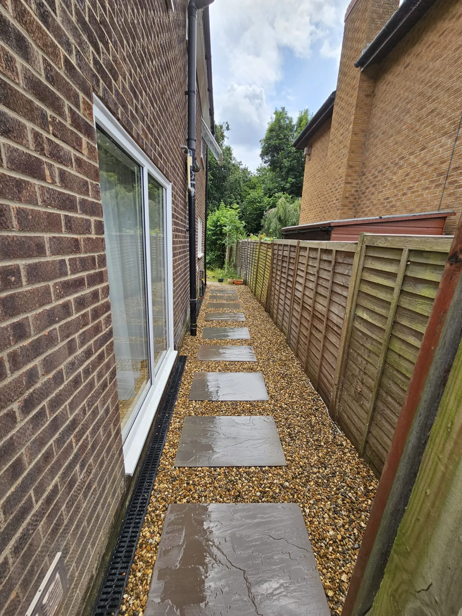 A black channel drain installed between a residential brick wall and a decorative gravel path with stepping stones, managing surface water.