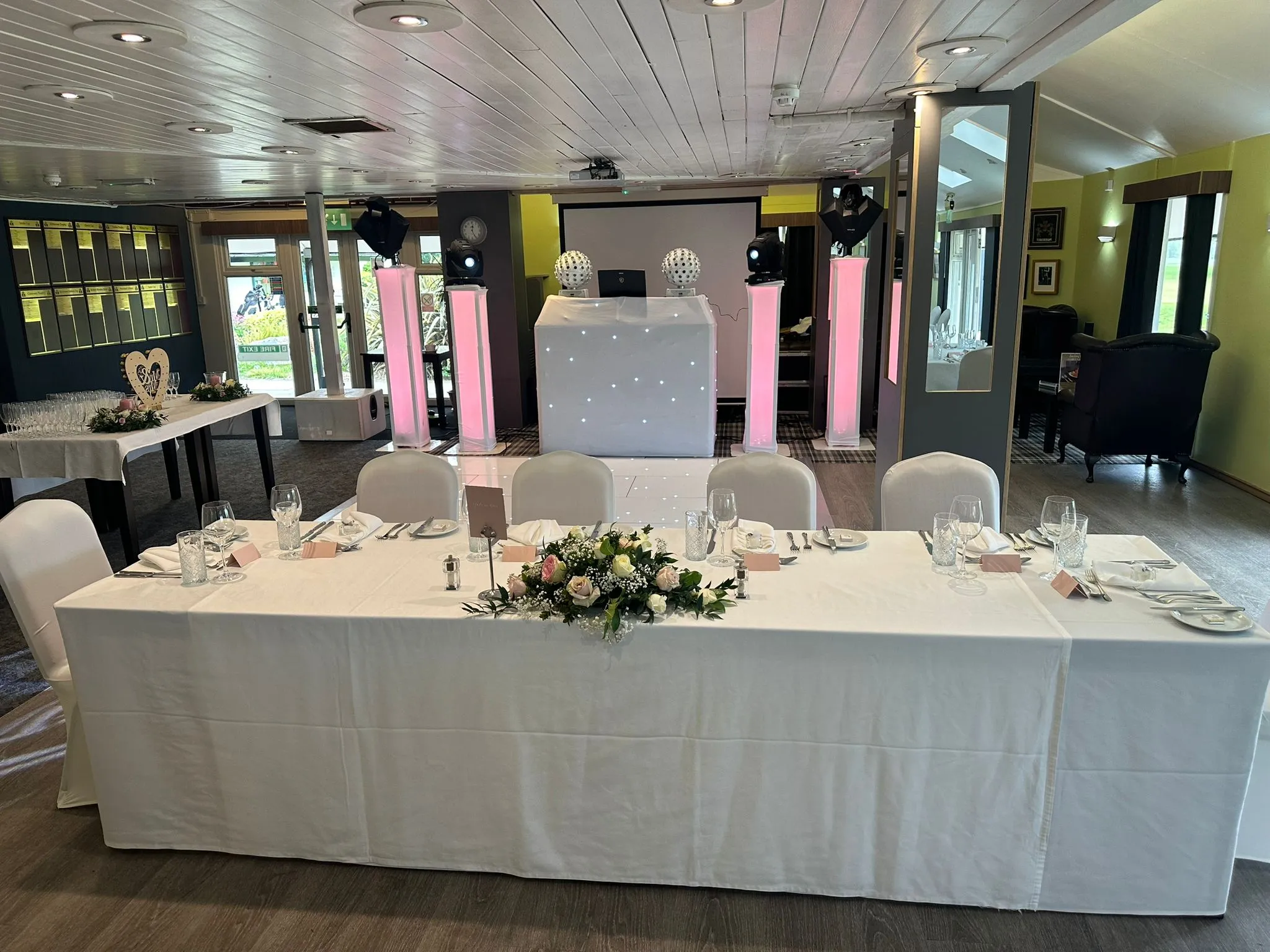 A stylish wedding reception room setup with a sparkling white DJ booth, pink uplighting on pillars, a large screen, and a reflective white starlit dance floor, alongside a long head table decorated with floral arrangements and place settings.
