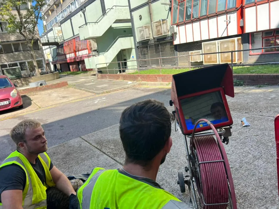 Two drainage technicians in high-visibility vests operating a red CCTV drain survey camera unit with a monitor displaying an internal view of a pipe.