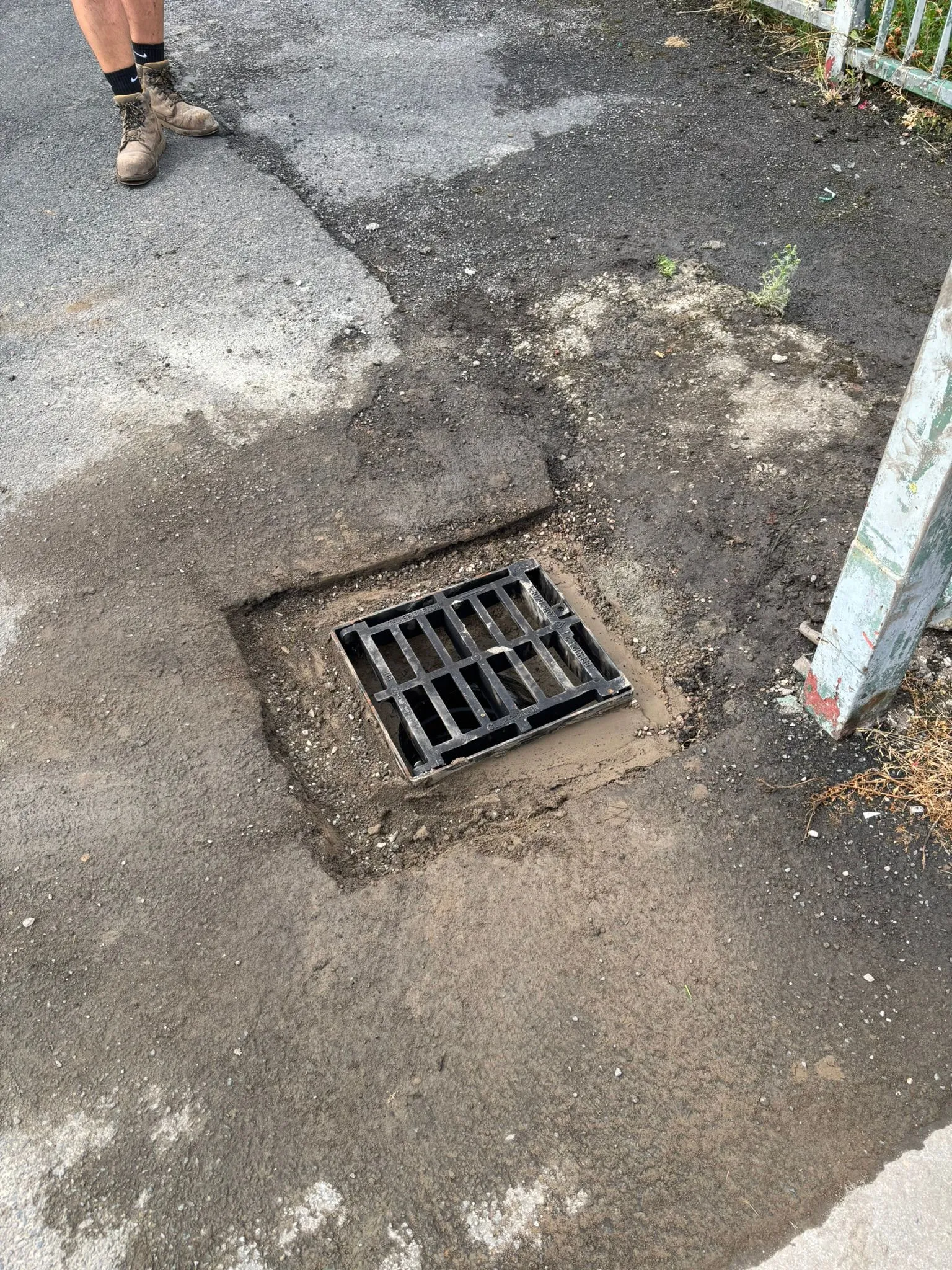 A drain grate being installed or repaired, surrounded by wet soil and gravel, with a worker's boots nearby.