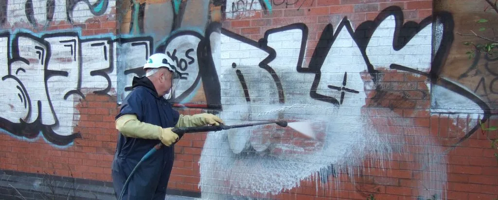 A worker using a high-pressure water jet to clean a graffiti-covered brick wall