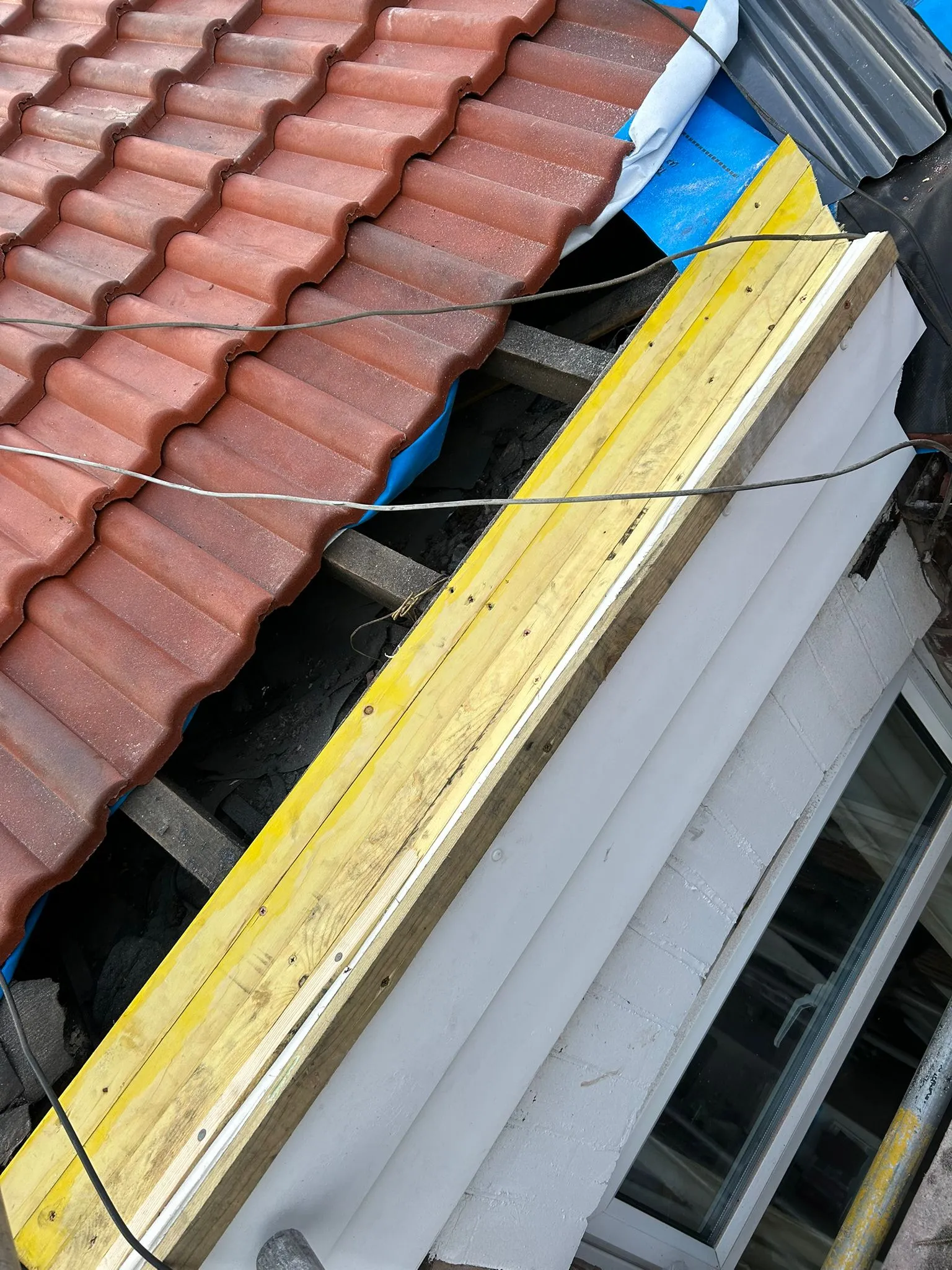 Detail of a pitched roof installation or repair in progress, featuring red tiles, blue waterproof membrane, and new wooden framing.