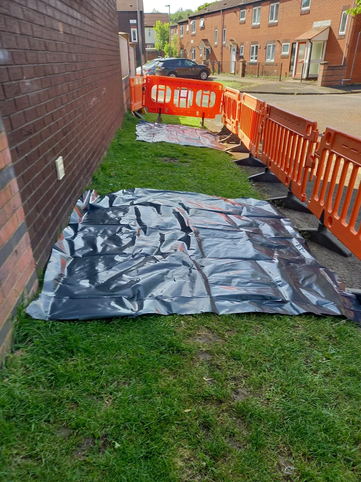 Orange safety barriers and black protective sheeting laid out on a grassy area next to a brick building and residential street, showing preparation for drainage work.