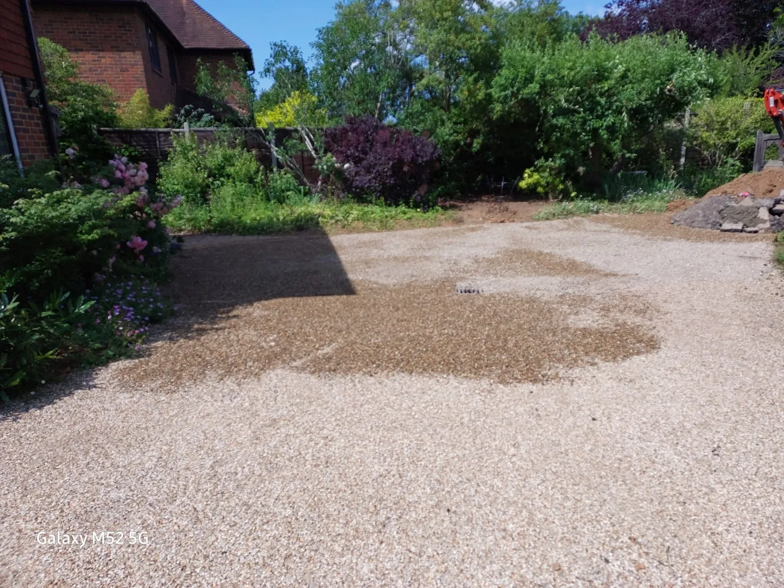 A drain grate in a gravel driveway, surrounded by a patch of darker, possibly damp, gravel, with a residential house and garden in the background.