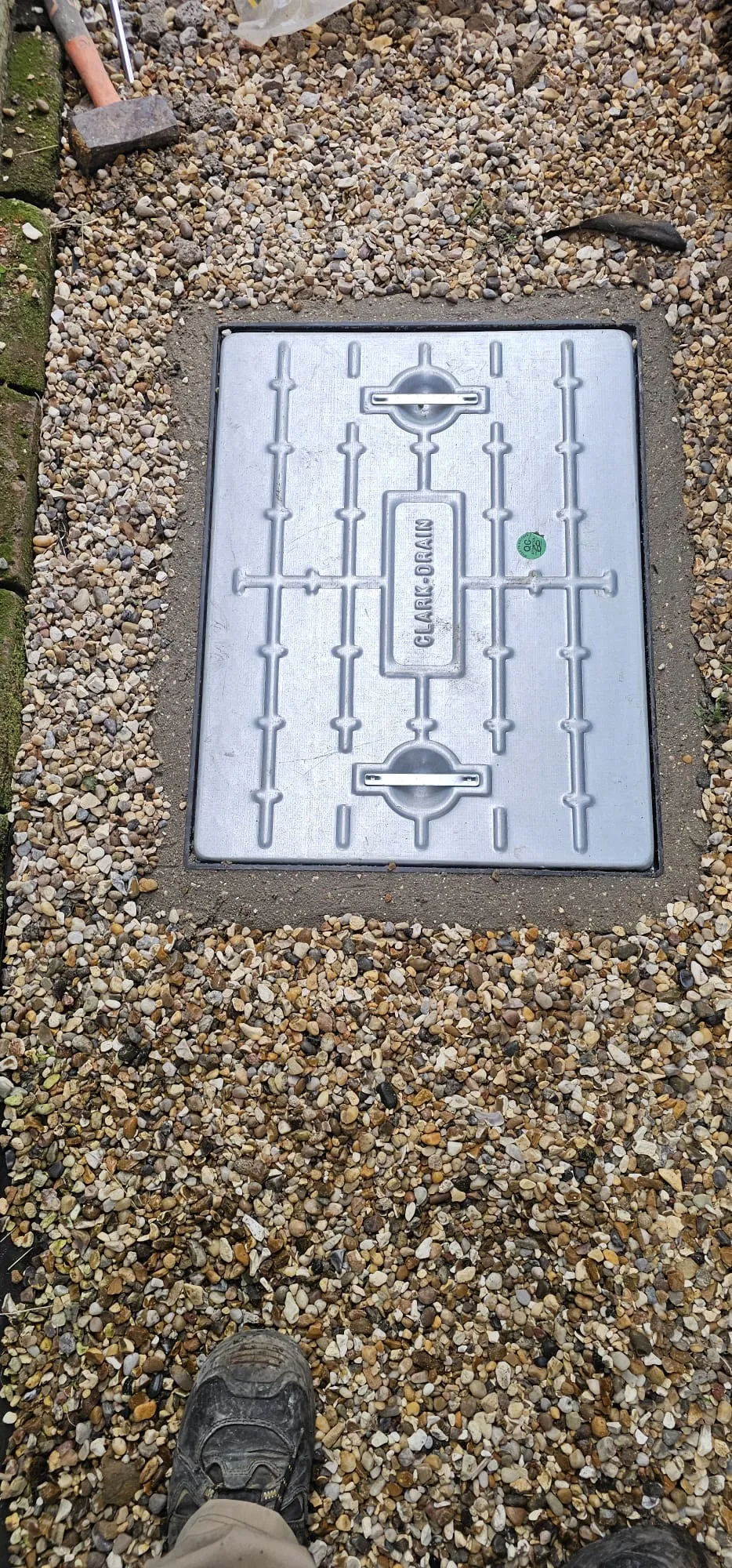 Metal Clark-Drain manhole cover set in gravel with work tools nearby.