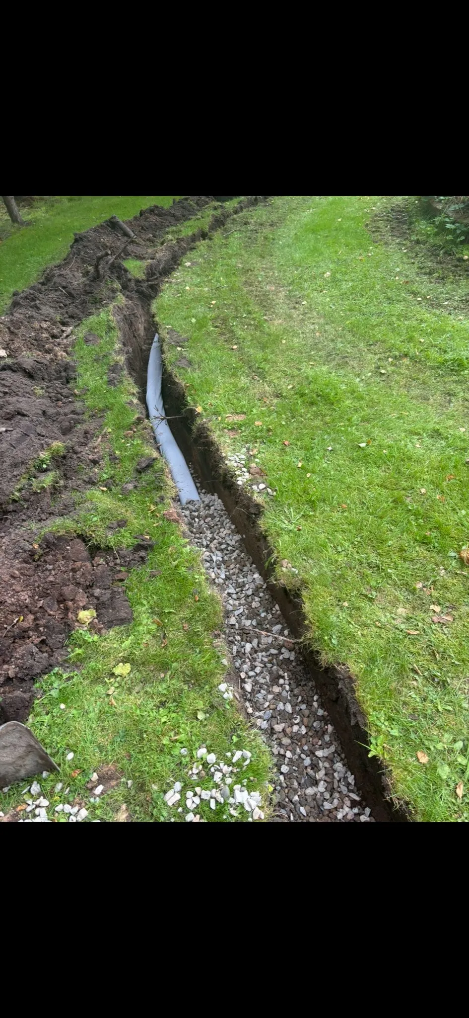 A dug trench in a green lawn with a grey drainage pipe on gravel, illustrating drain excavation and installation work.