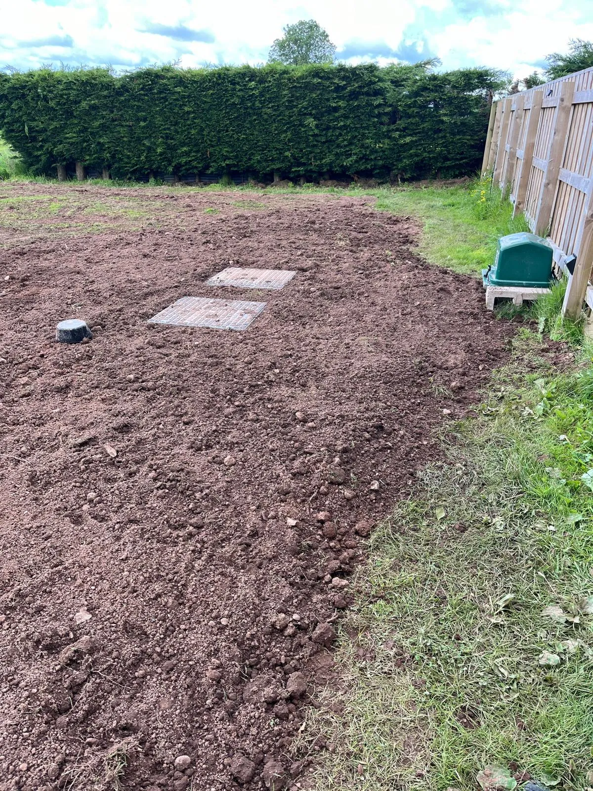 Newly prepared ground in a garden with visible drainage access covers and a green pump station or sewage treatment plant.