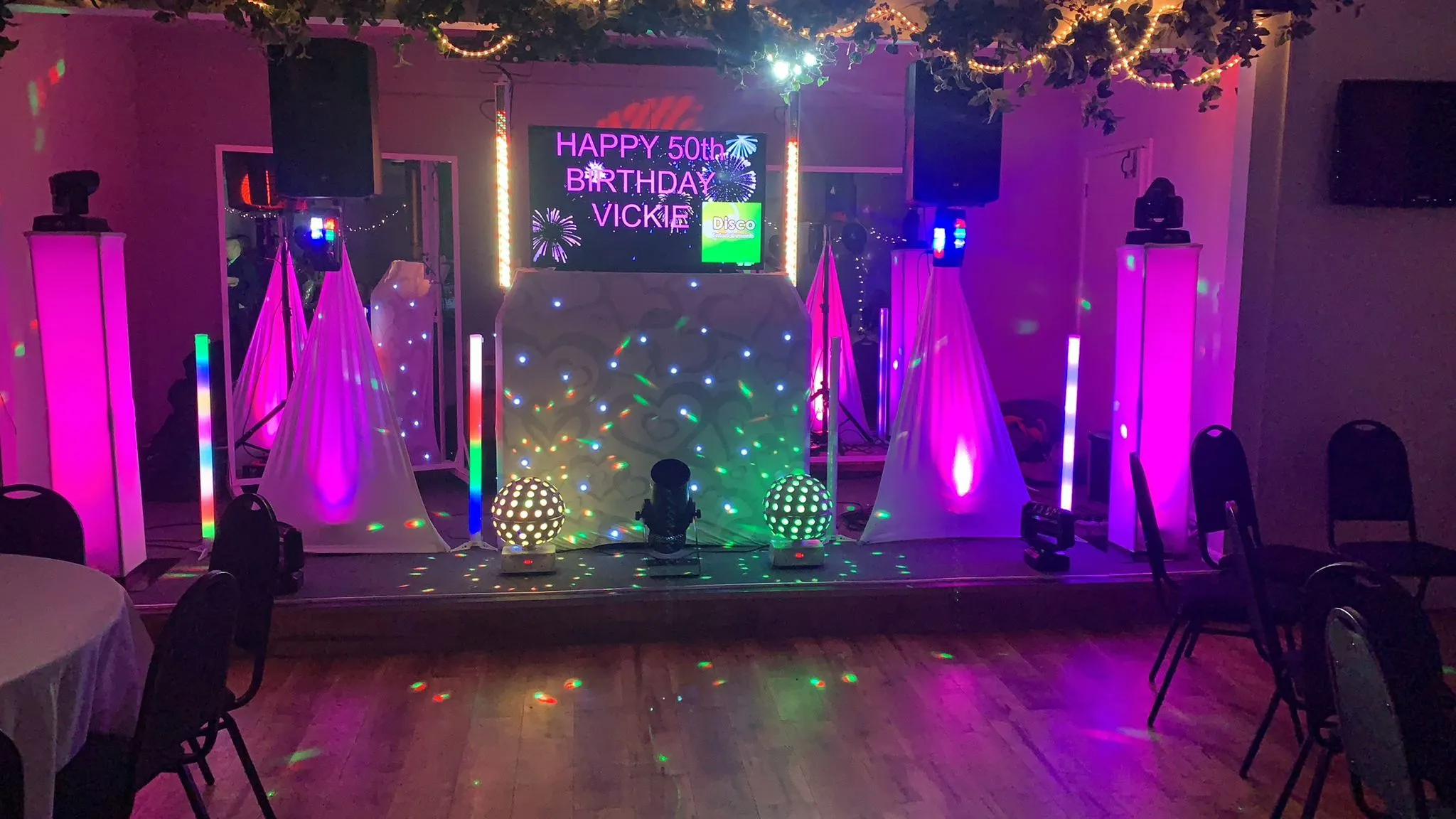 A professional DJ setup by Disco Entertainers for a 50th birthday party, featuring a custom screen displaying 'HAPPY 50th BIRTHDAY VICKIE', surrounded by vibrant purple and multi-coloured lights.