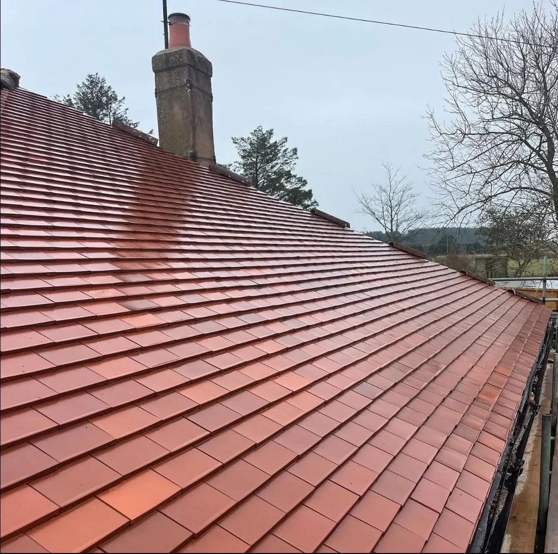 A close-up view of a recently completed pitched roof installation, showcasing new terracotta-coloured plain tiles and a traditional brick chimney stack against a cloudy sky.