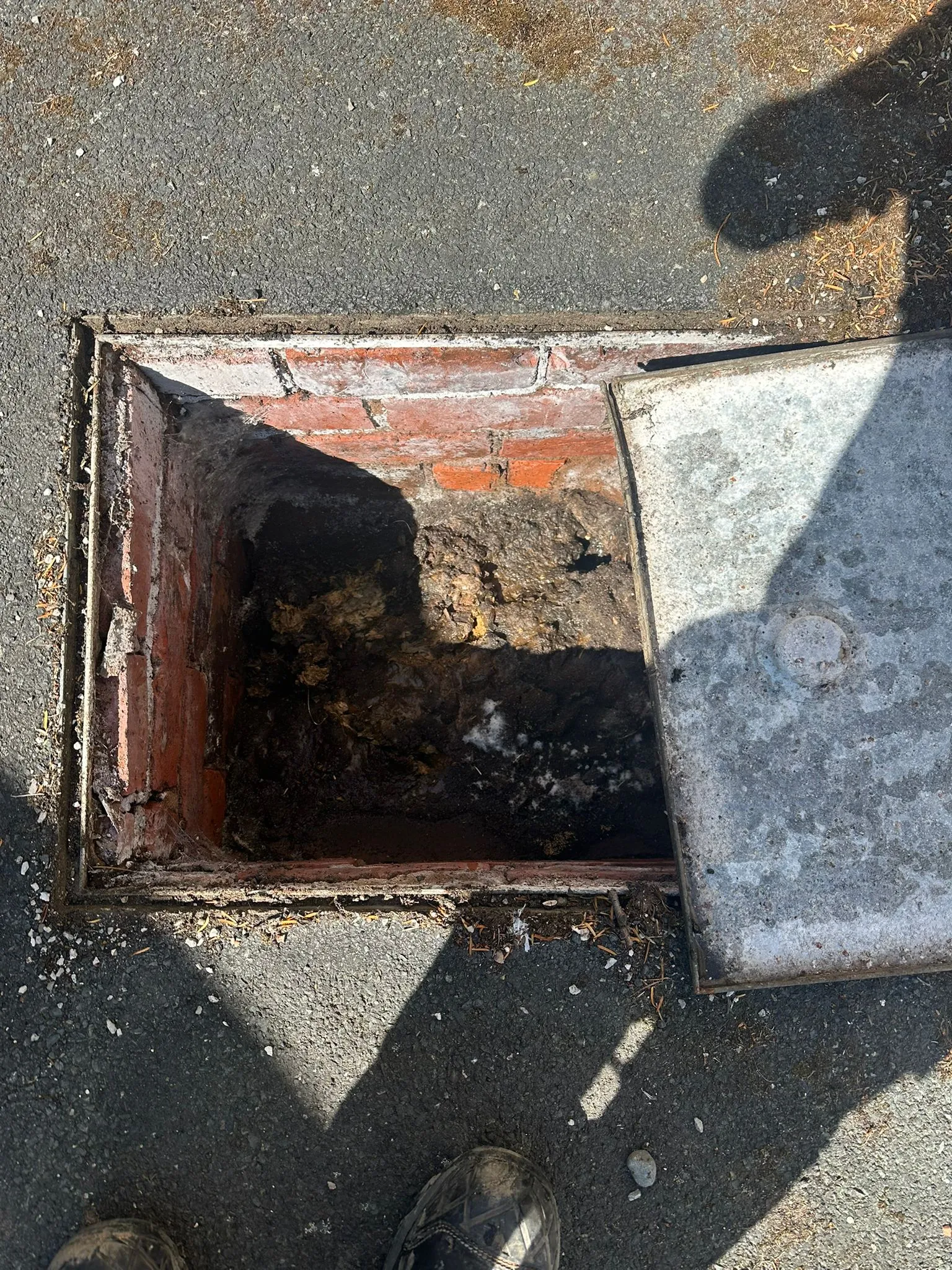 Open manhole showing a blocked drain with heavy sludge and debris inside, on an asphalt surface.