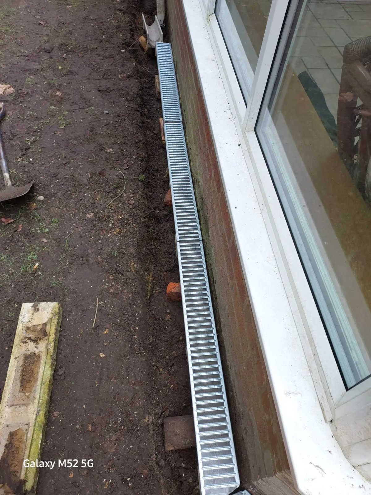 A newly installed linear channel drain with a metal grate, running along a brick wall next to windows, with muddy ground and a spade nearby, indicating drainage work in progress.