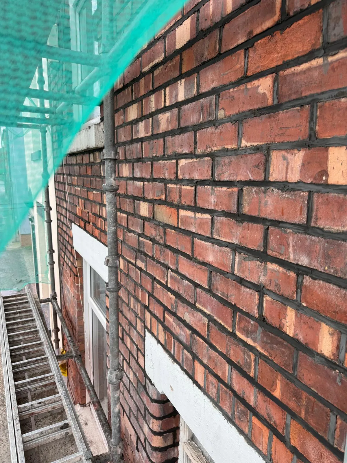 Close-up of a recently repointed red brick wall with dark mortar, featuring scaffolding and green netting.