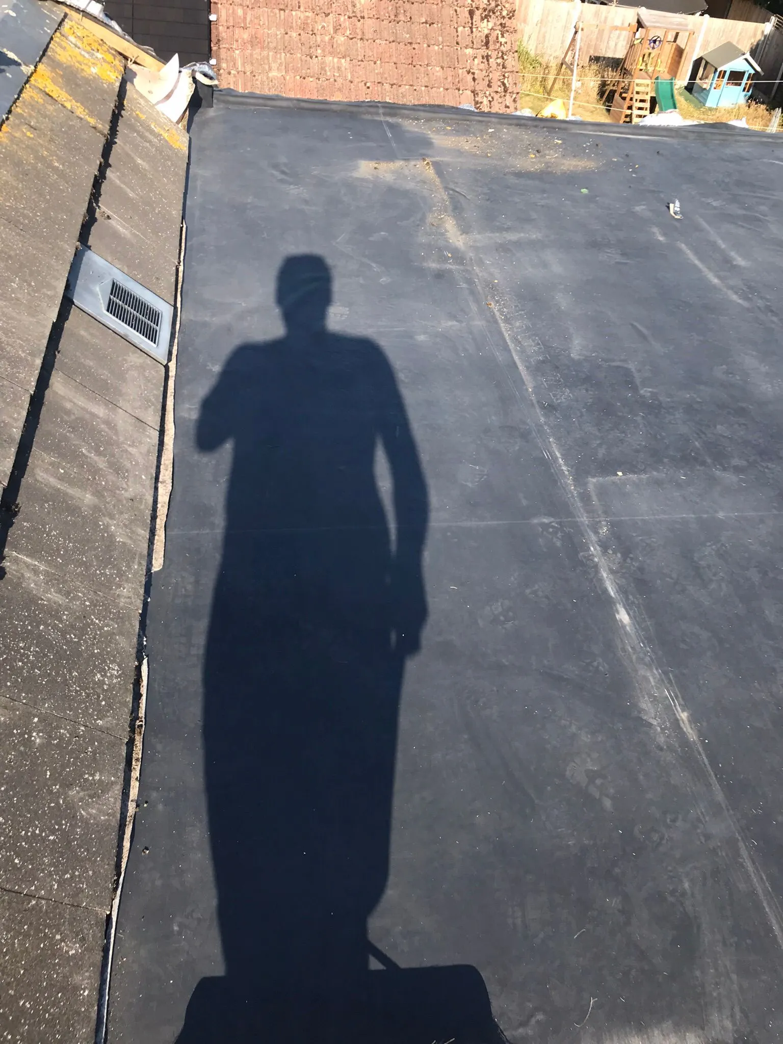 A roofer's view across a flat roof adjacent to a pitched tiled roof, showing both roofing types and a roof vent in a sunny residential setting.