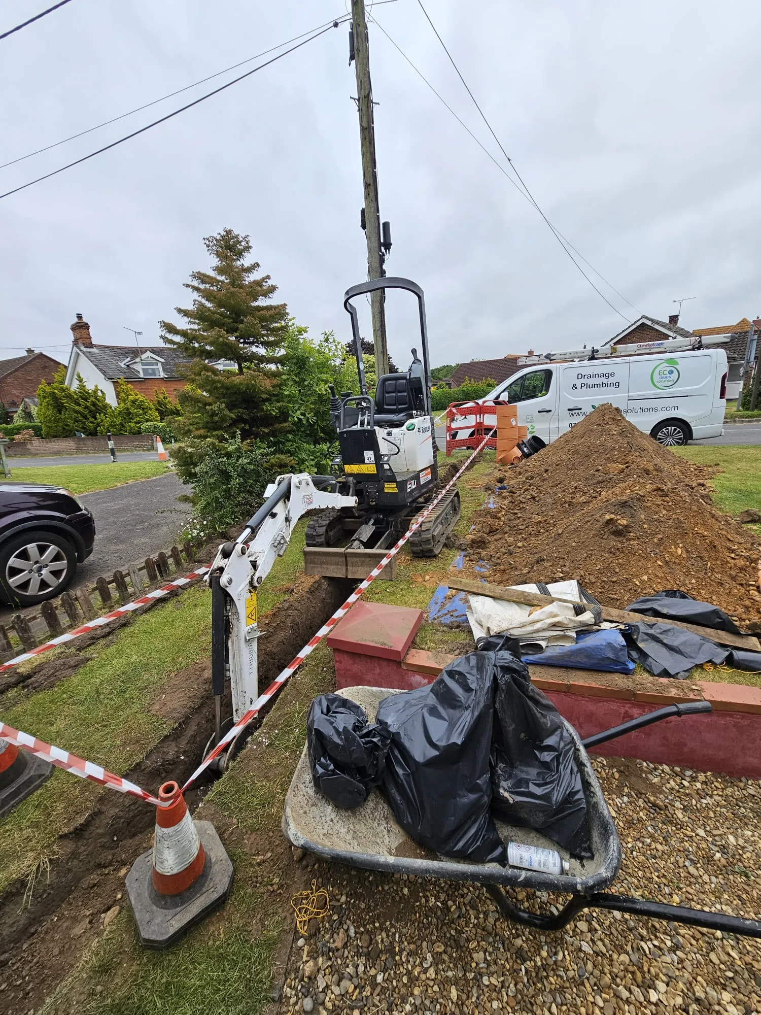 A small white excavator digging a trench in a grassy verge, marked with red and white safety tape and traffic cones. A large pile of soil is next to the trench, and a white Eco Drain Solutions van is visible further down the street.