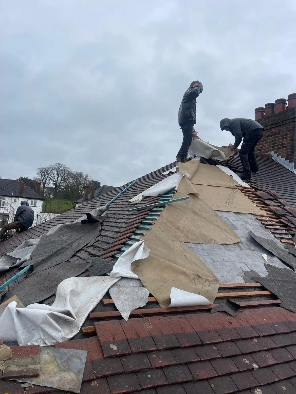 Roofers replacing a pitched tile roof, showing exposed underlayment, battens, and removed tiles.