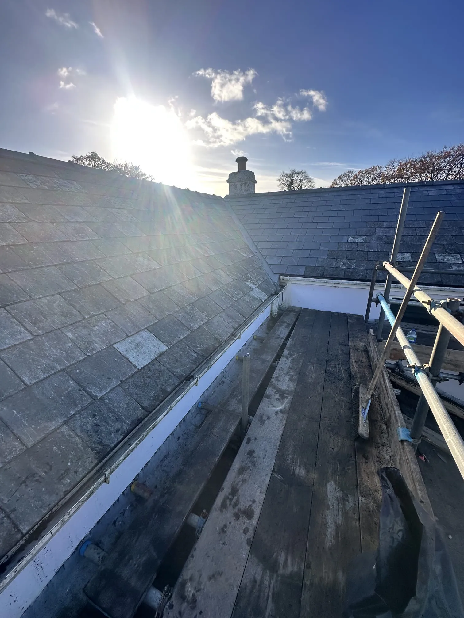 Slate roof with scaffolding, a chimney, and bright sun, indicating roof work in progress.