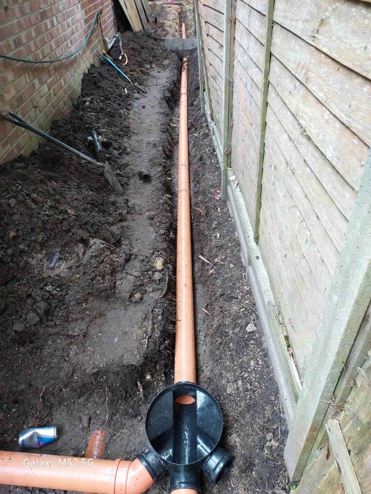 An open trench with new orange drainage pipes and a black inspection chamber fitting, alongside a brick wall and wooden fence.