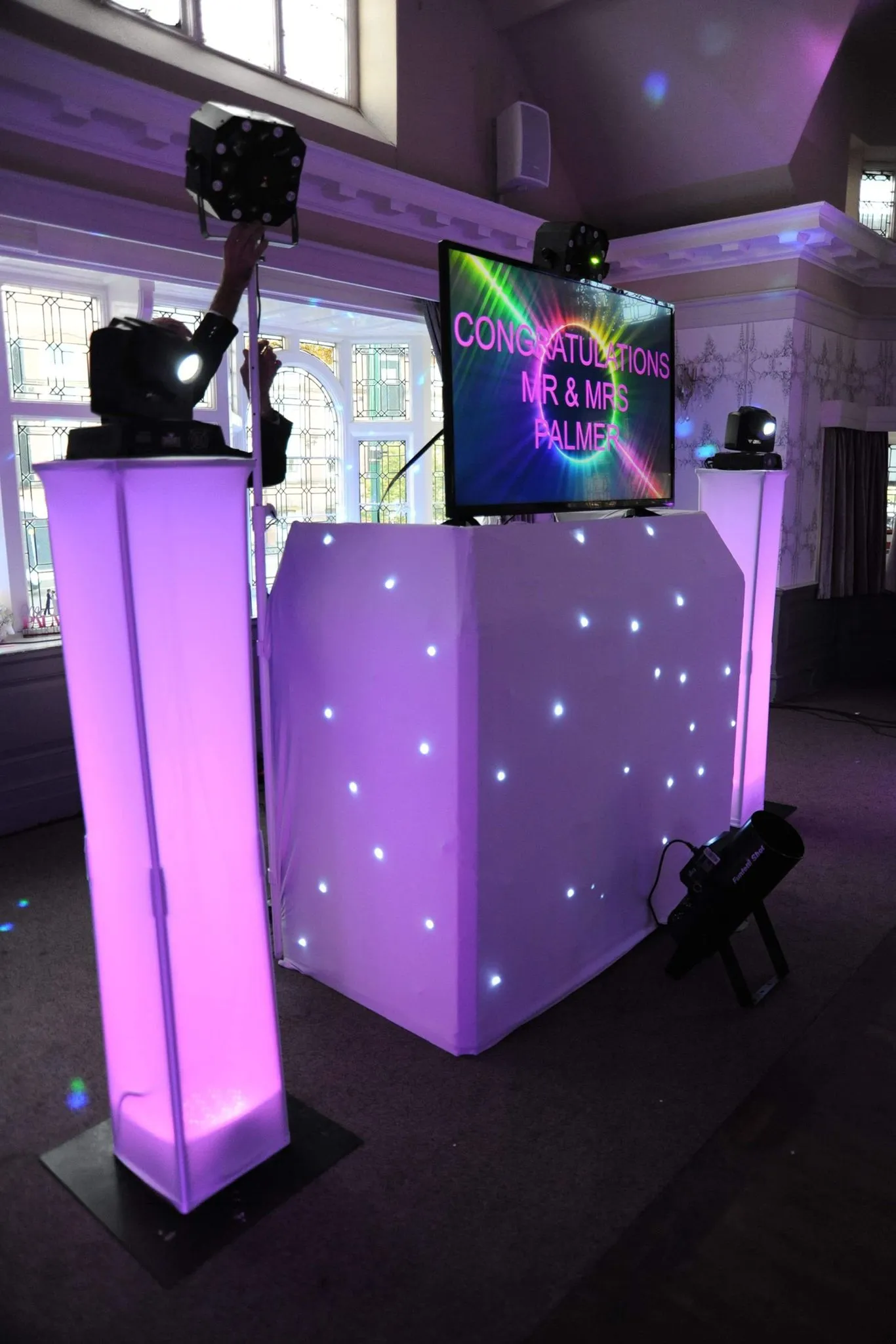 A professional wedding DJ setup with purple ambient lighting, a starlit white DJ booth, and a large screen displaying 'Congratulations Mr & Mrs Palmer'.