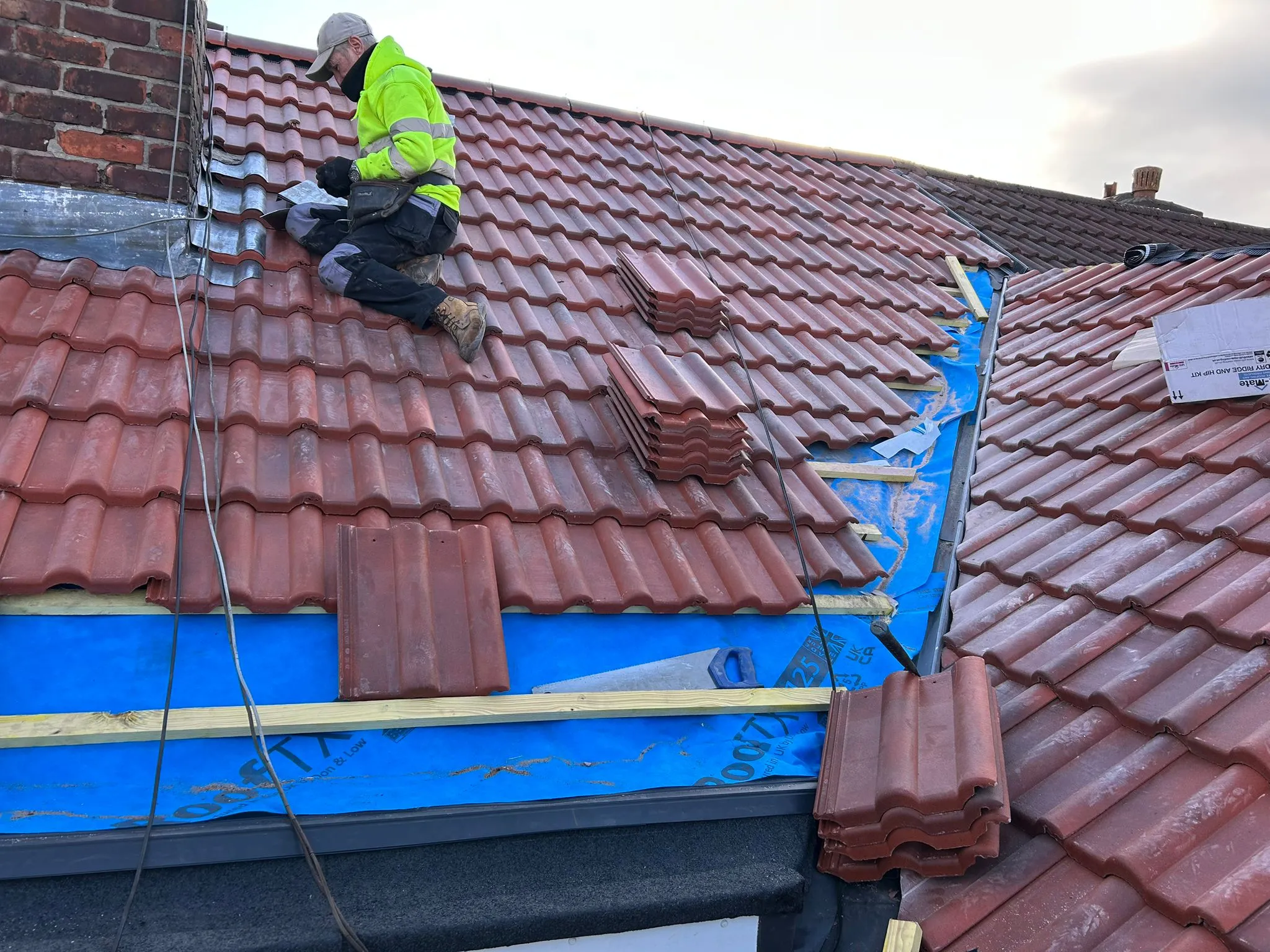 Professional roofer installing red interlocking tiles and lead flashing on a pitched roof, with blue waterproof underlay visible beneath the new tiles.