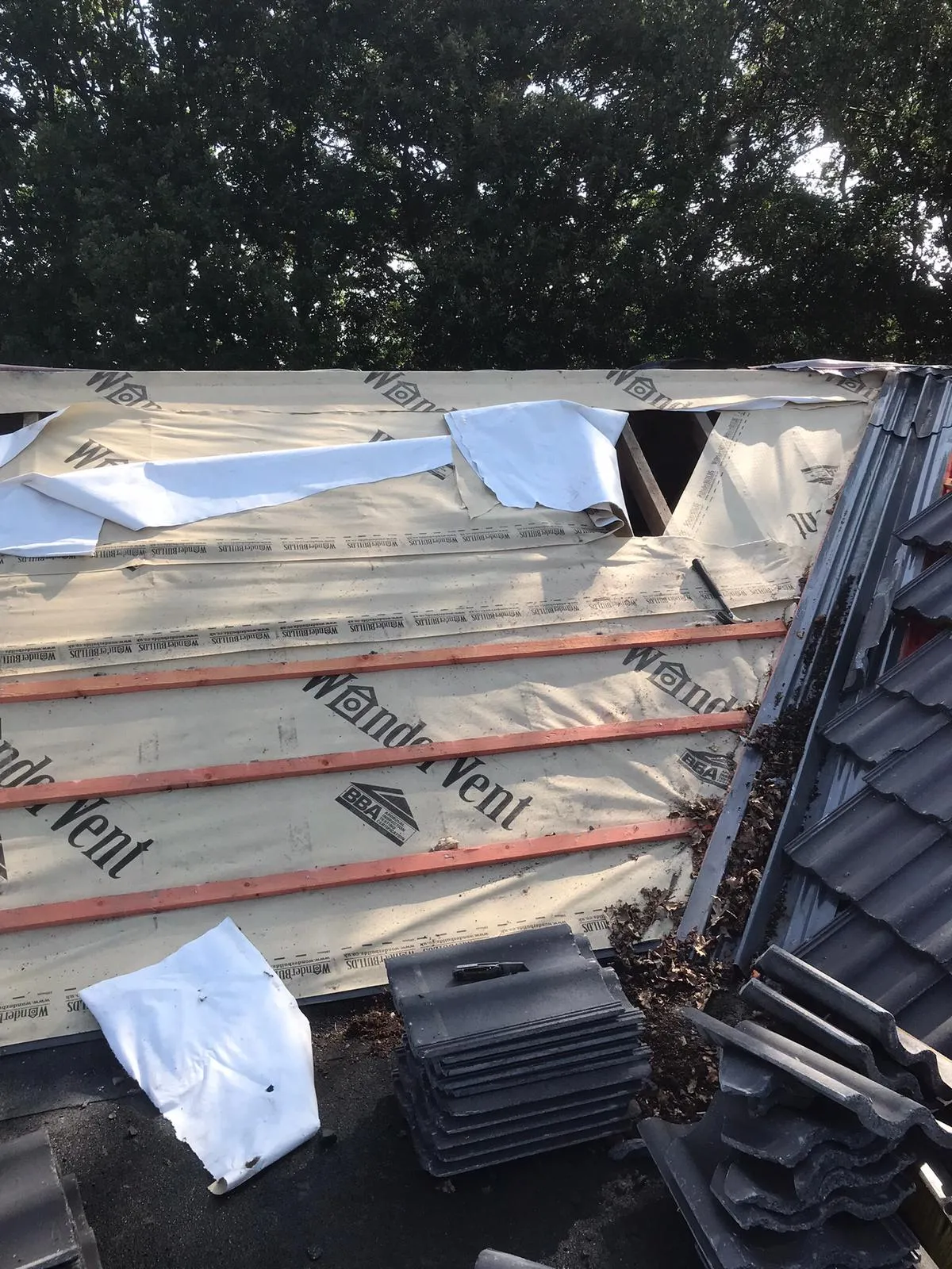 An active roofing project with a beige waterproof membrane and wooden battens installed over the roof structure, with new dark grey roof tiles stacked nearby, ready for placement.