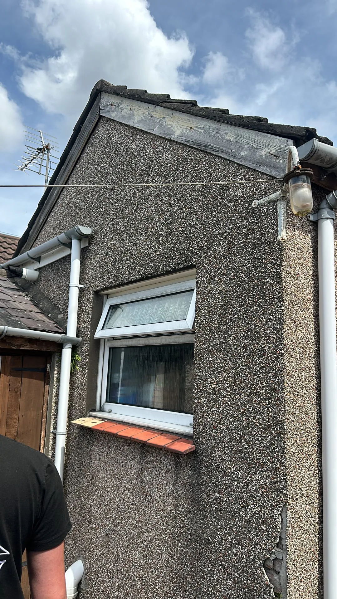Side of a house with worn timber fascias and a pitched roof, showing the roofline and an open window.