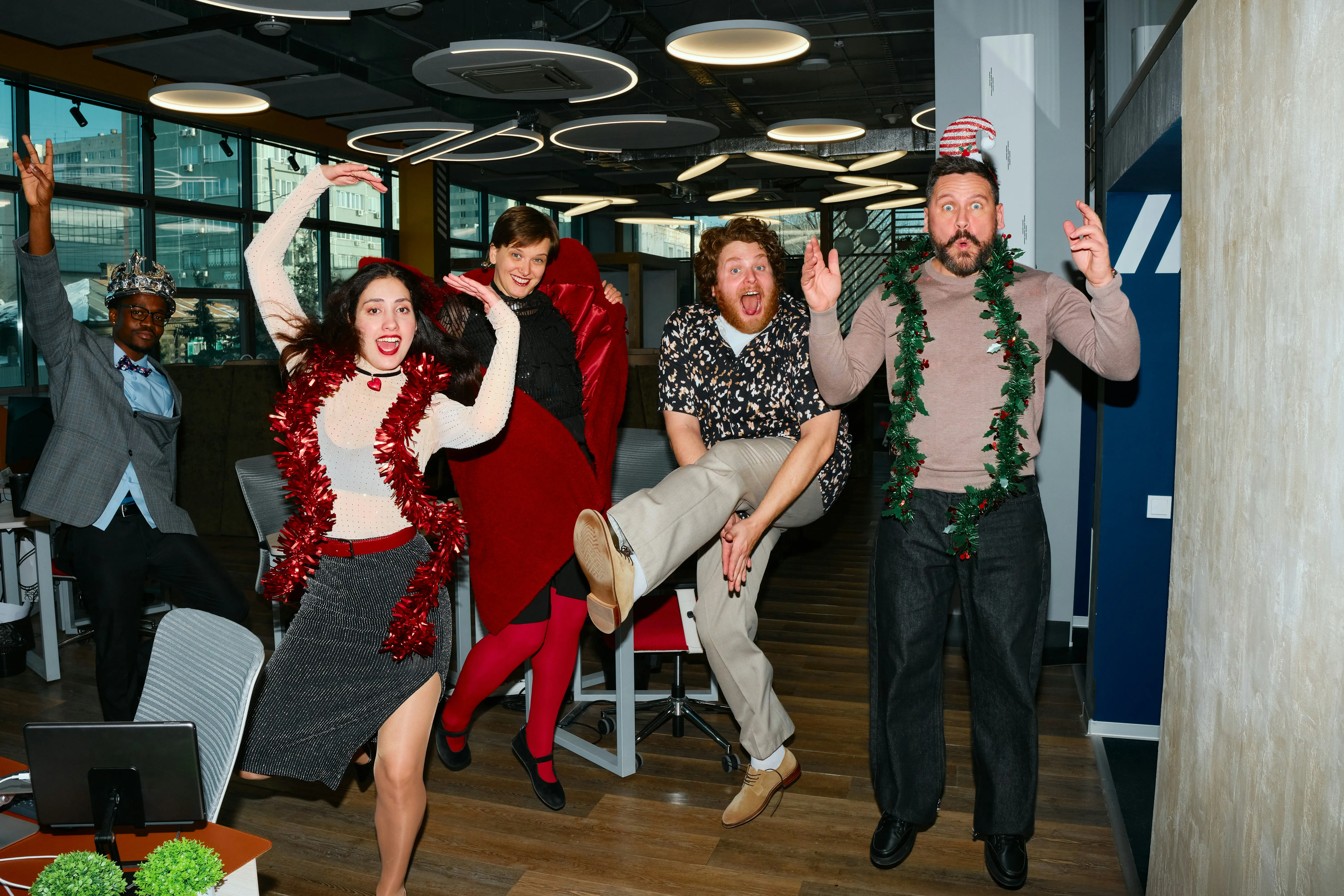 Five diverse people, dressed in party attire with tinsel and hats, jumping and posing energetically in a modern office space, celebrating with joy.