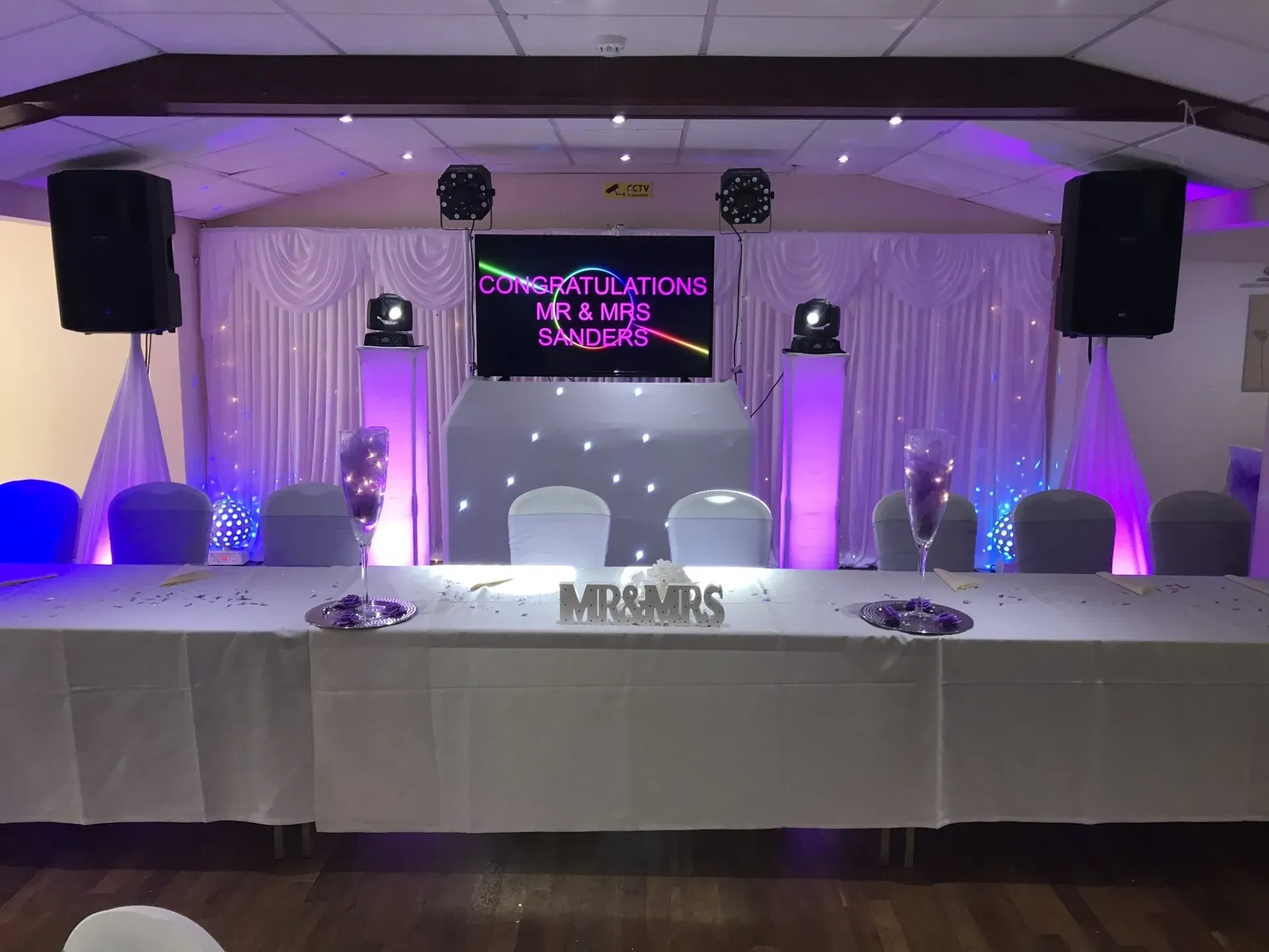 Wedding top table with DJ setup, screen showing congratulations message, purple uplighting, speakers, and white backdrop
