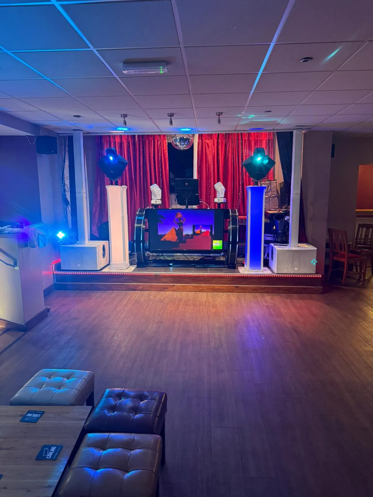 An impressive event DJ setup from Disco Entertainers, showcasing a large central screen with music video playback, bright blue and white moving head lights, LED uplighting, a reflective disco ball, and a professional sound system on a raised stage with red backdrop curtains.